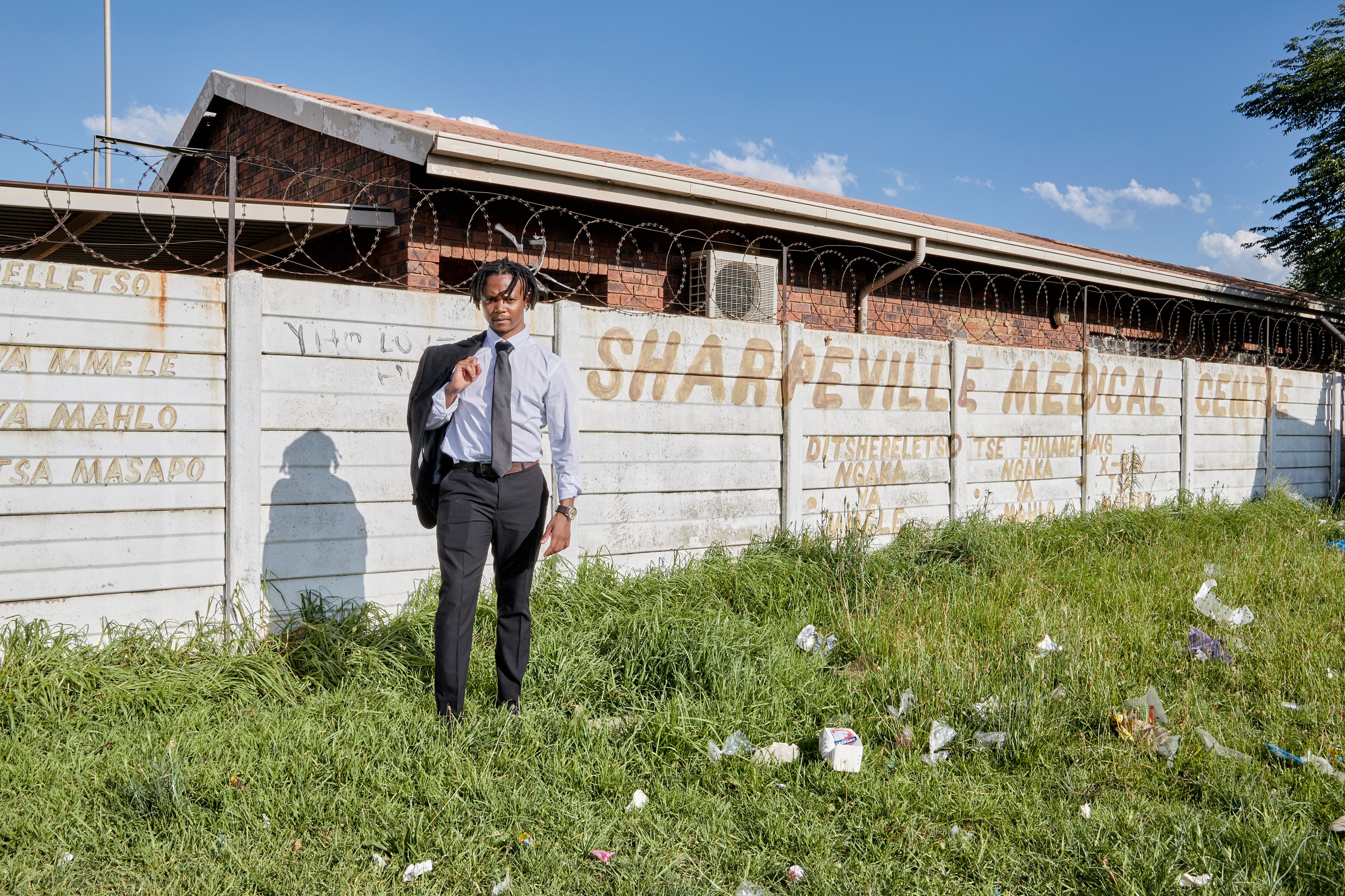 A man in business attire stands in overgrown, littered grass in front of a concrete wall with "Sharpeville Medical Centre" written on it and razor wire on top.