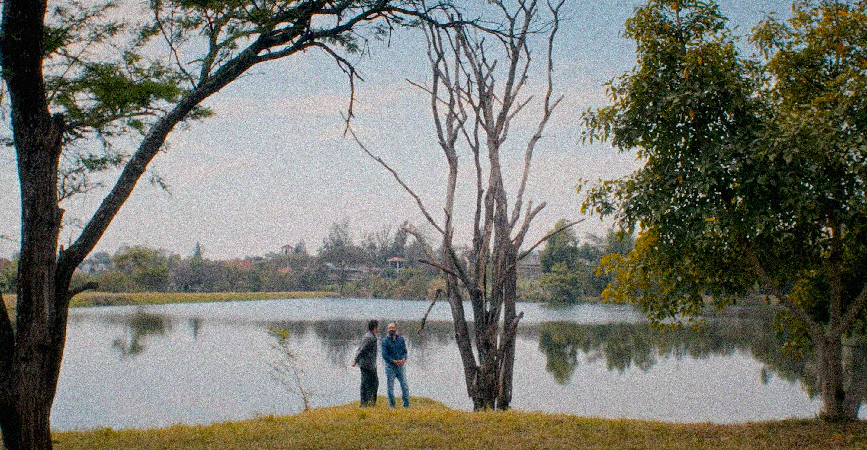 Two men stand on a grassy bank beside a lake, surrounded by trees.