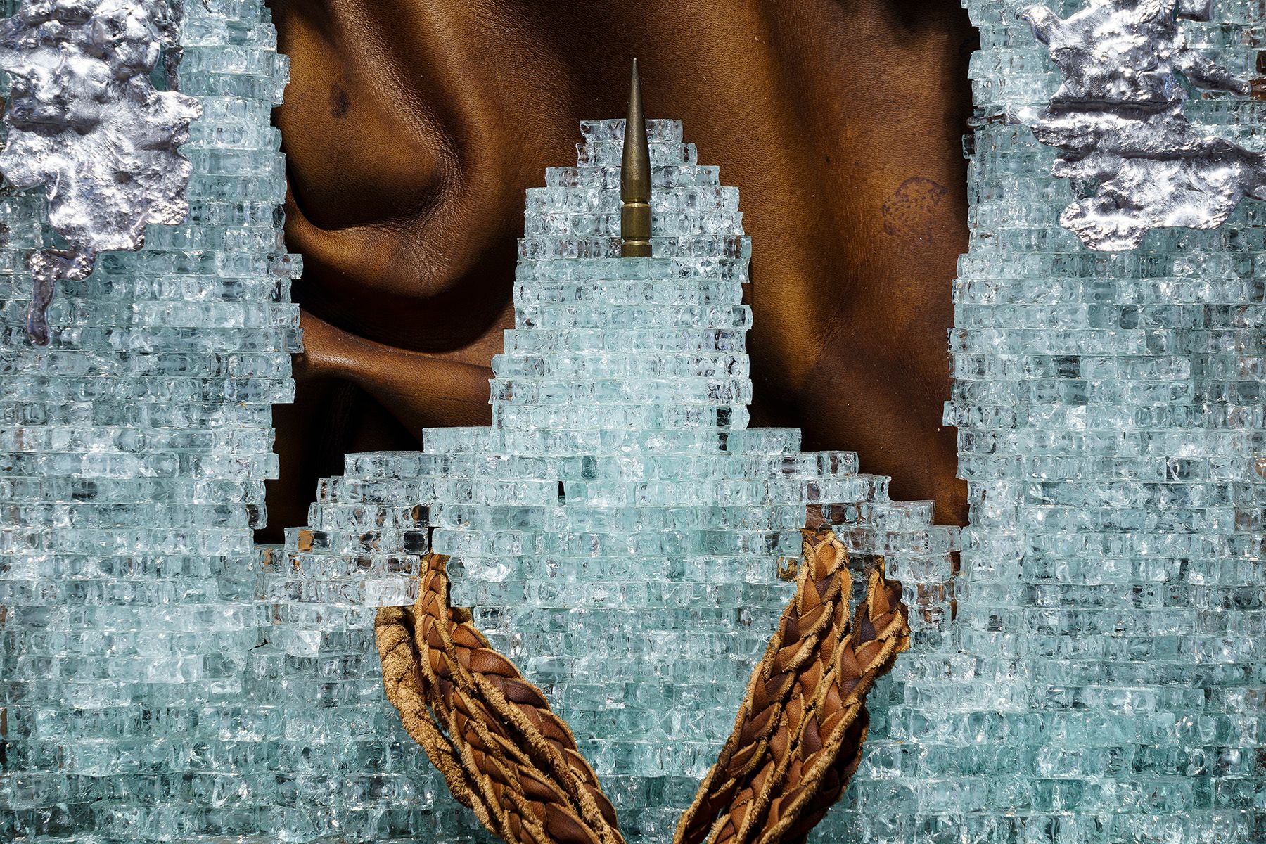 A brown, textured surface is visible through a jagged opening in a field of light blue glass shards, with a bullet at the top and two braided brown leather strands at the bottom.