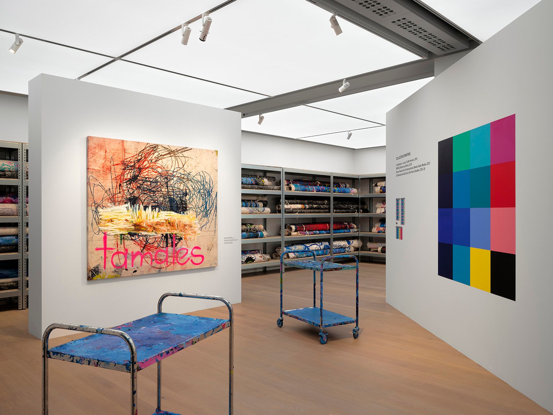 An exhibition space featuring an abstract painting with "tamales" written in pink, a colorful grid of squares, shelves of rolled textiles, and two paint-splattered utility carts.