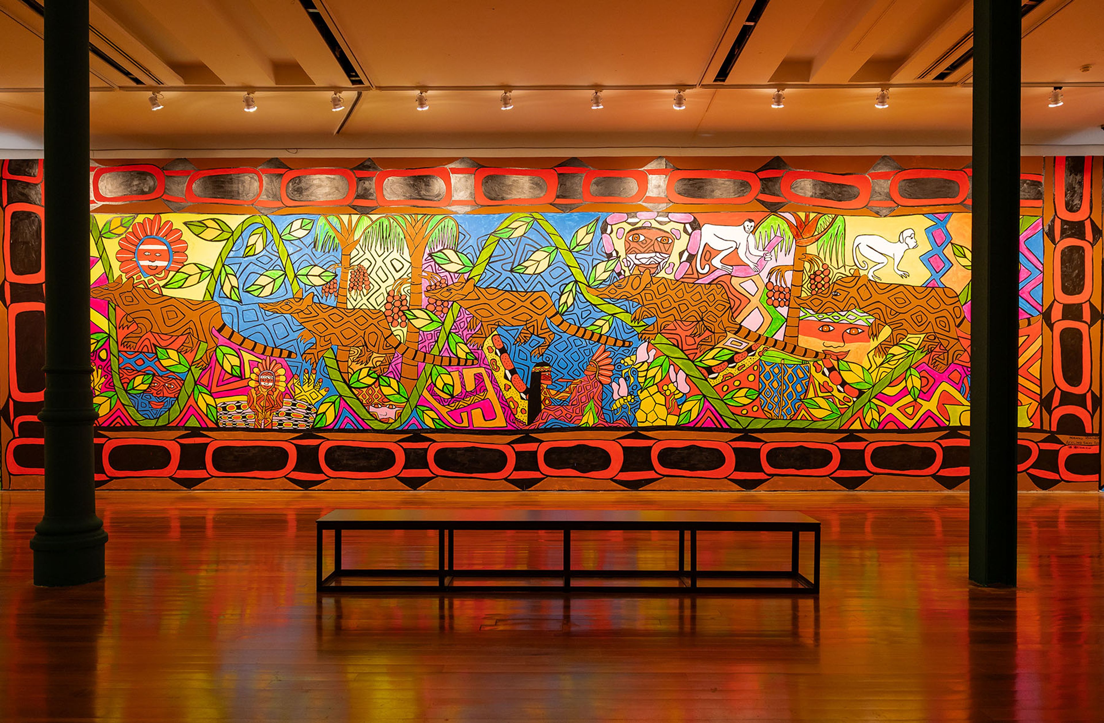 A large, vibrant mural featuring stylized jaguars, monkeys, and jungle flora in bright colors fills an art gallery wall, with a black bench on a reflective wooden floor.