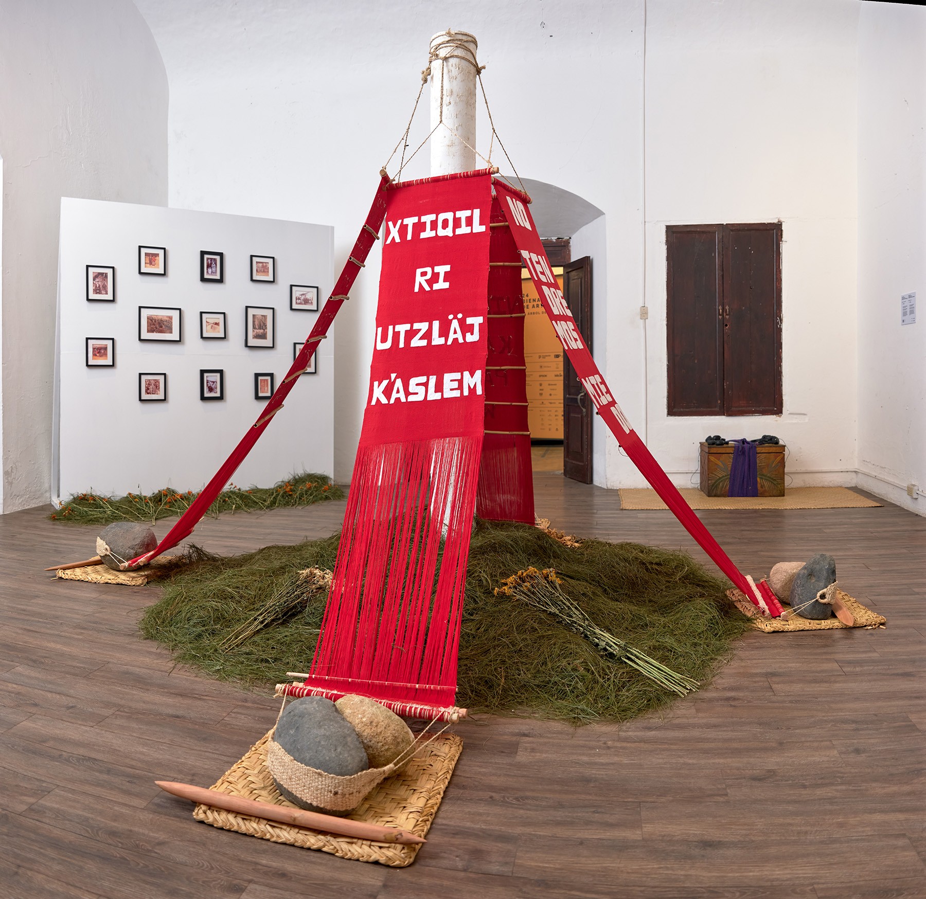 An art installation with red fabric banners displaying text hanging from a central pillar, anchored by stones on a bed of green plant material, in a gallery setting.
