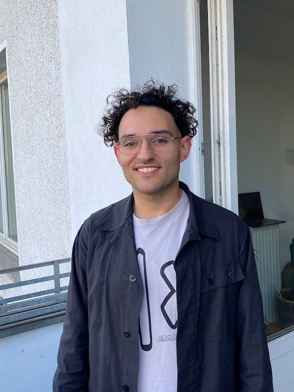 A smiling man with curly hair and glasses wears a black jacket over a light t-shirt.