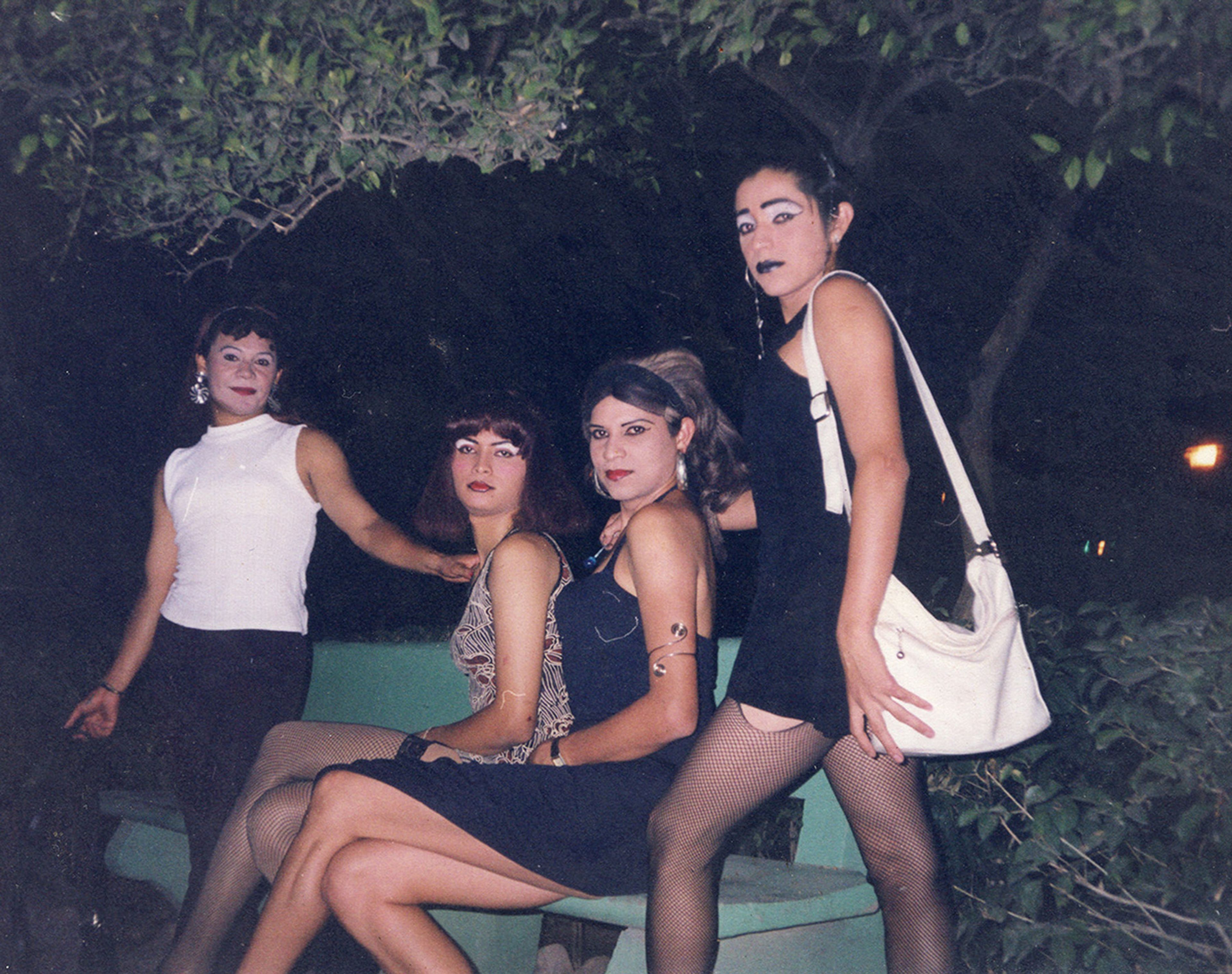 Four individuals in feminine attire and makeup pose outdoors at night, three seated on a bench and one standing.