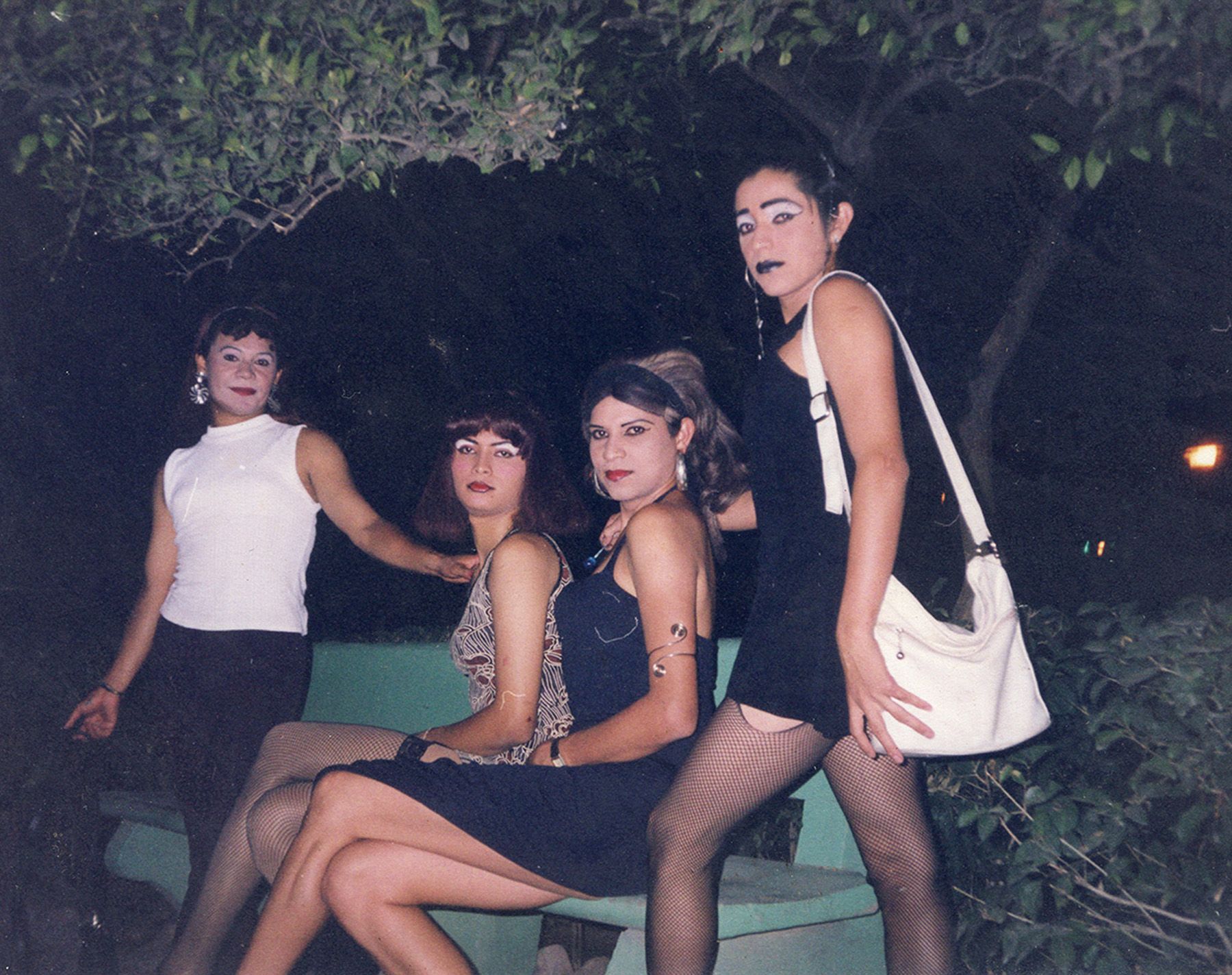 Four individuals in feminine attire and makeup pose outdoors at night, three seated on a bench and one standing.