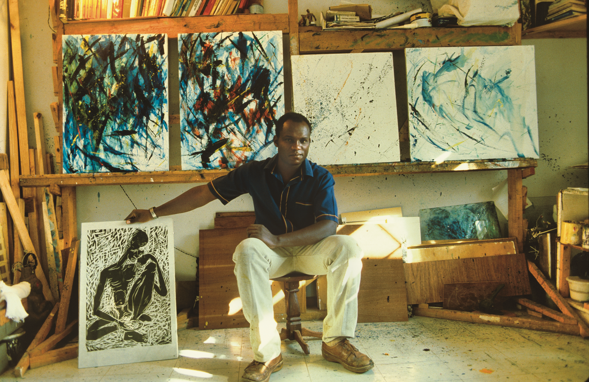A Black artist sits in his studio, holding a black and white print, with several abstract paintings behind him.