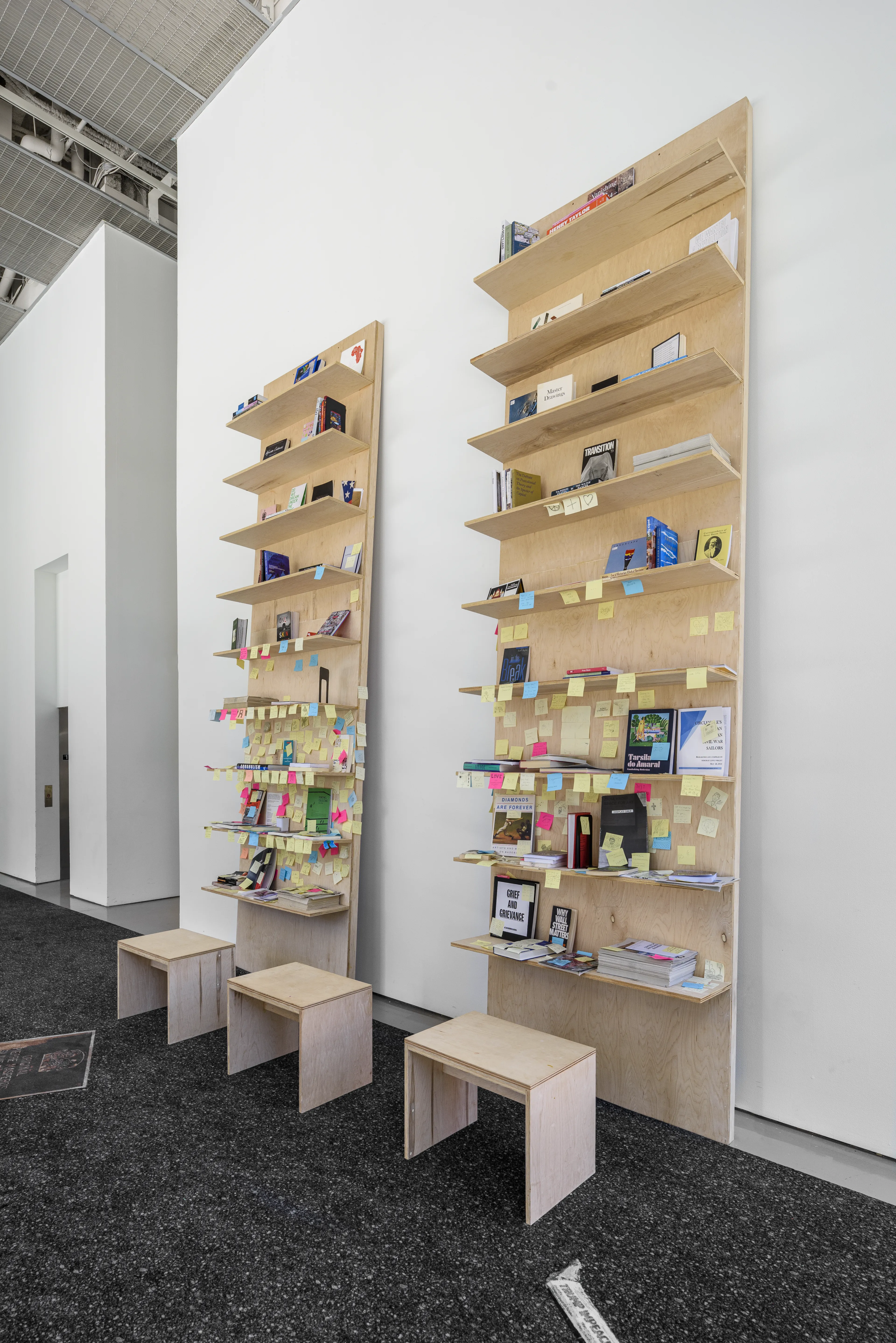 Two tall wooden bookshelves covered in books and colorful sticky notes, with matching stools, in a modern white room.