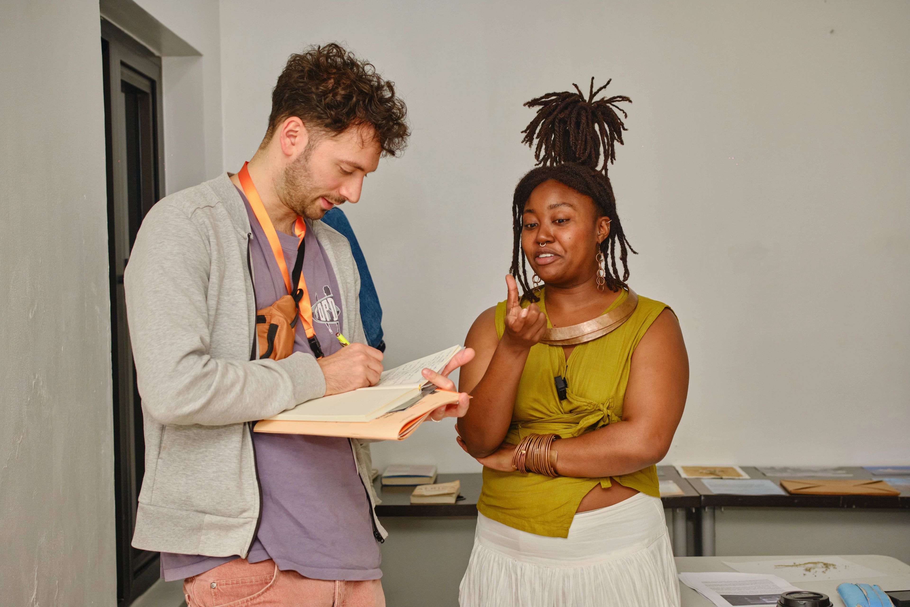 A man writes in a notebook while a woman with dreadlocks gestures and speaks to him.
