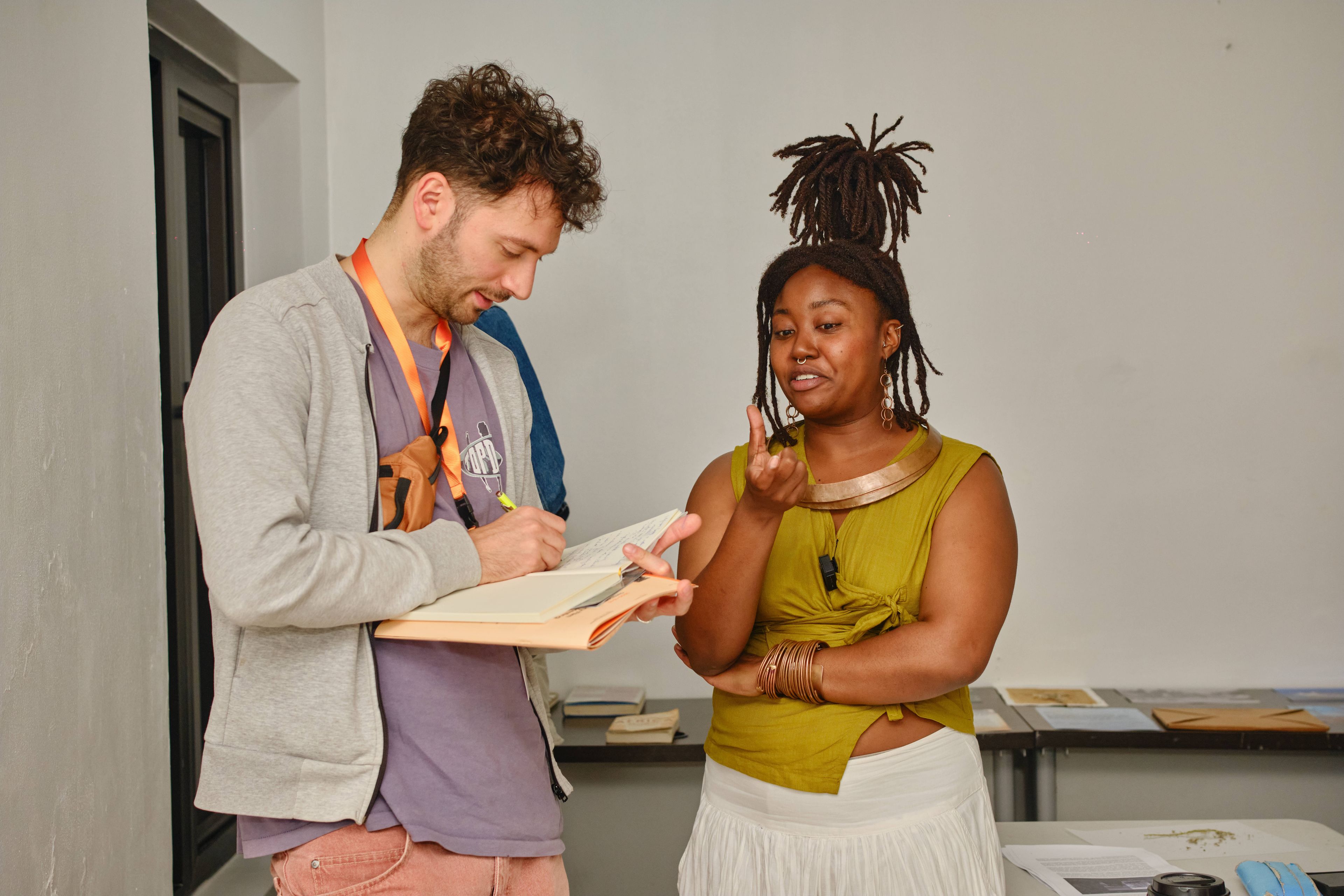 A man writes in a notebook while a woman with dreadlocks gestures and speaks to him.