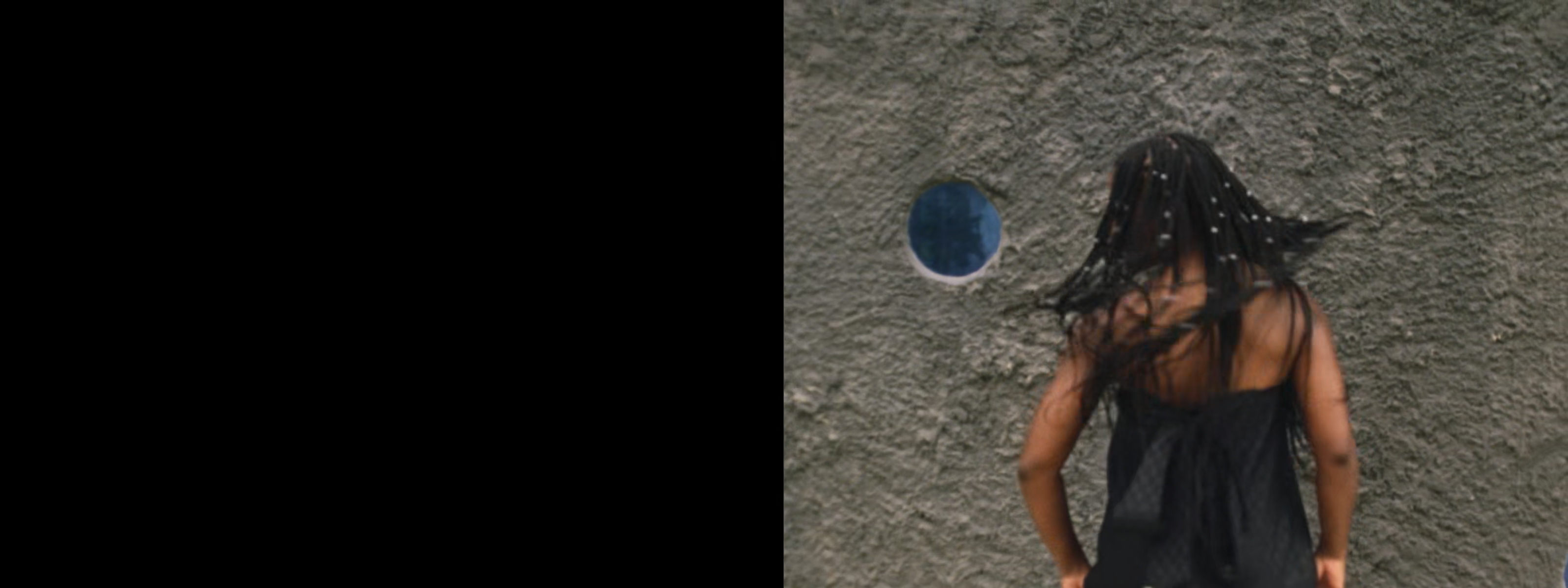 A person with flowing, beaded hair, seen from behind, stands against a textured gray wall with a circular blue element.