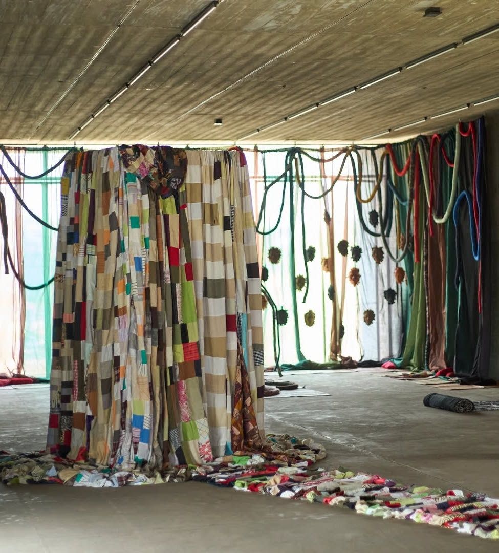 Art installation with a large patchwork quilt curtain, draped sheer fabrics, colorful ropes, and a fabric trail on a concrete floor.