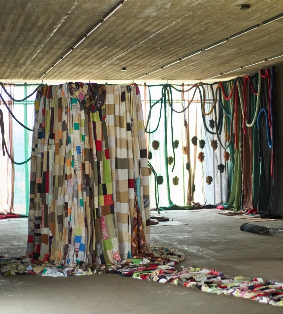 Art installation with a large patchwork quilt curtain, draped sheer fabrics, colorful ropes, and a fabric trail on a concrete floor.