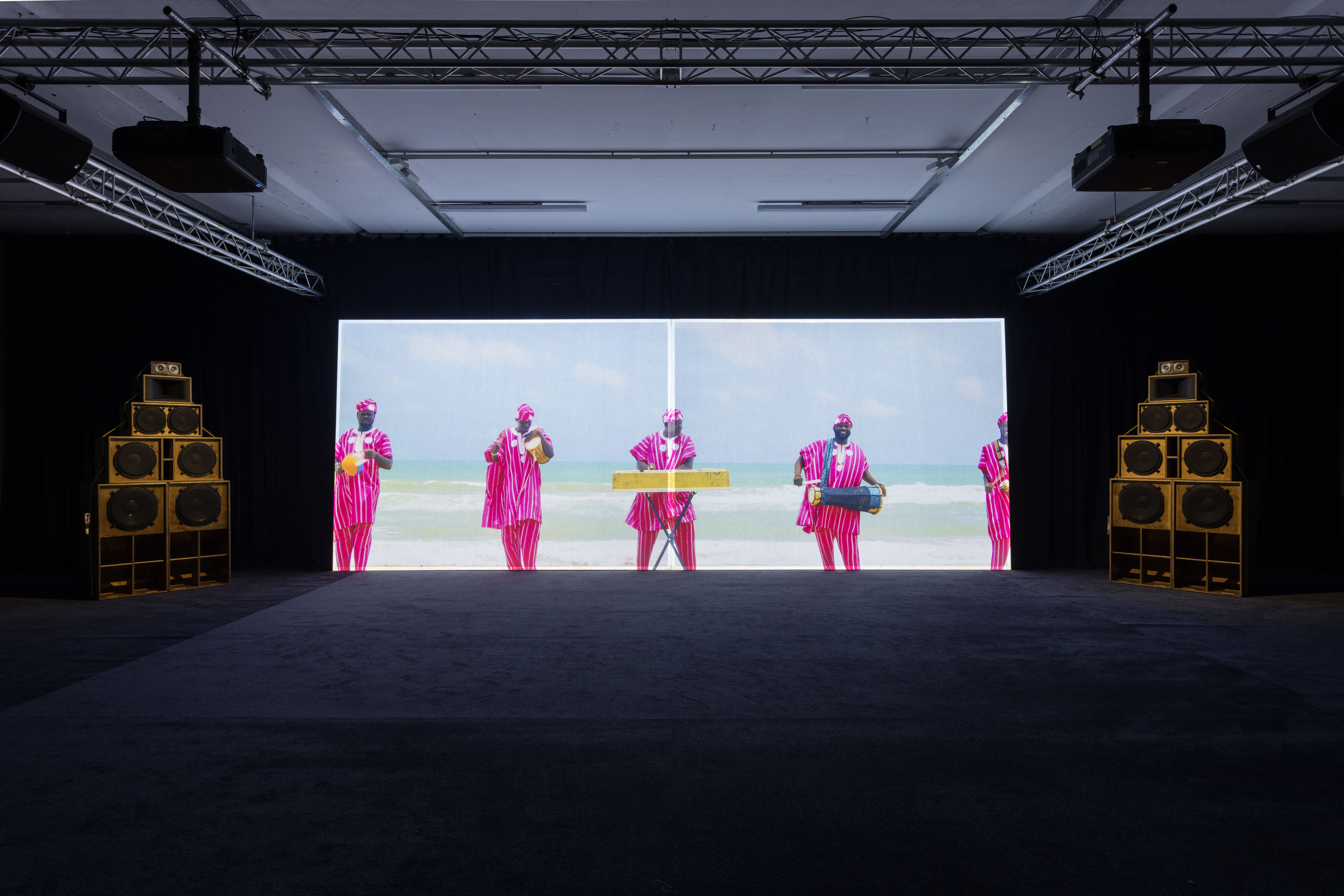 A dark exhibition room with a large video screen showing people in pink on a beach, flanked by two tall stacks of wooden speakers.