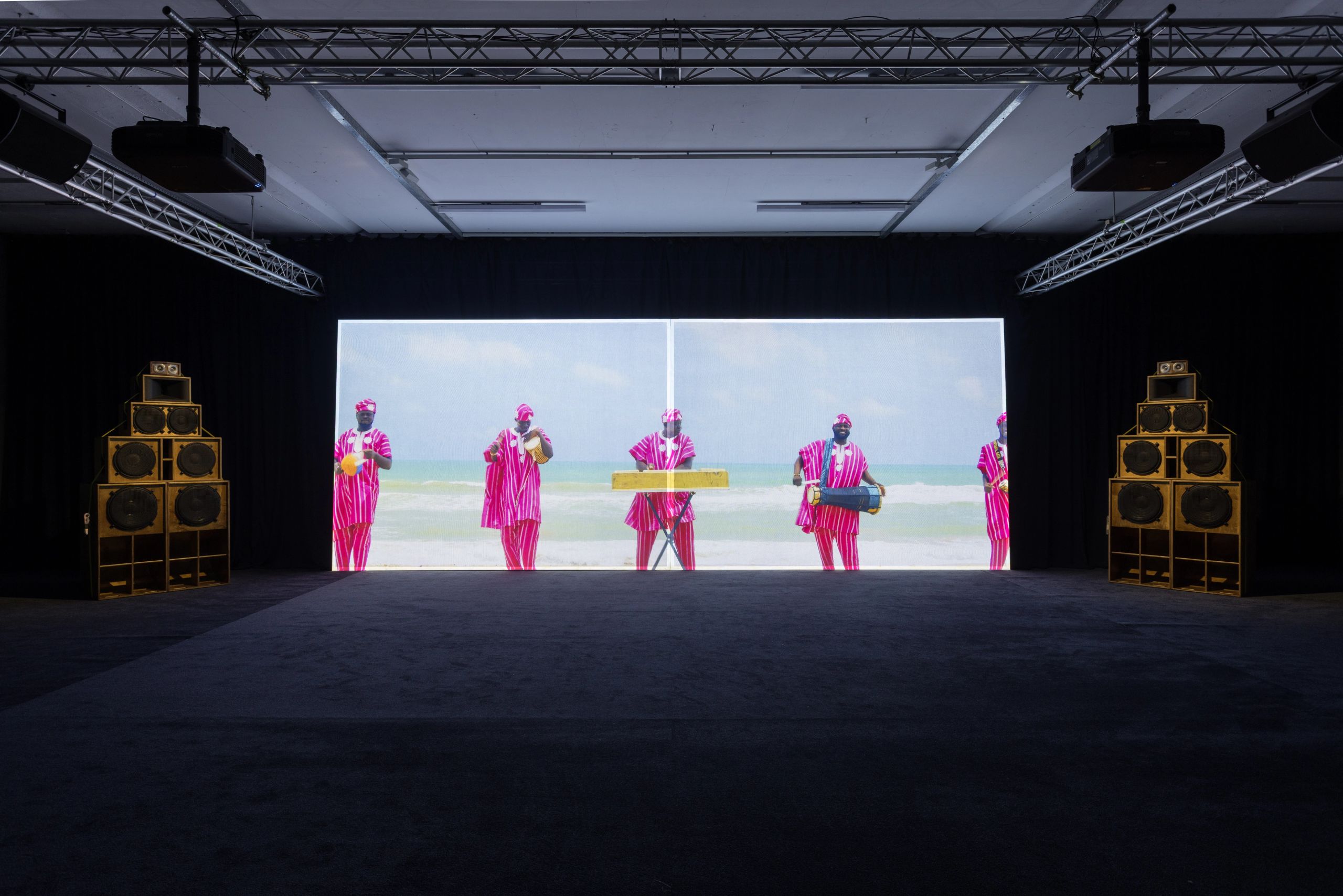 A dark exhibition room with a large video screen showing people in pink on a beach, flanked by two tall stacks of wooden speakers.
