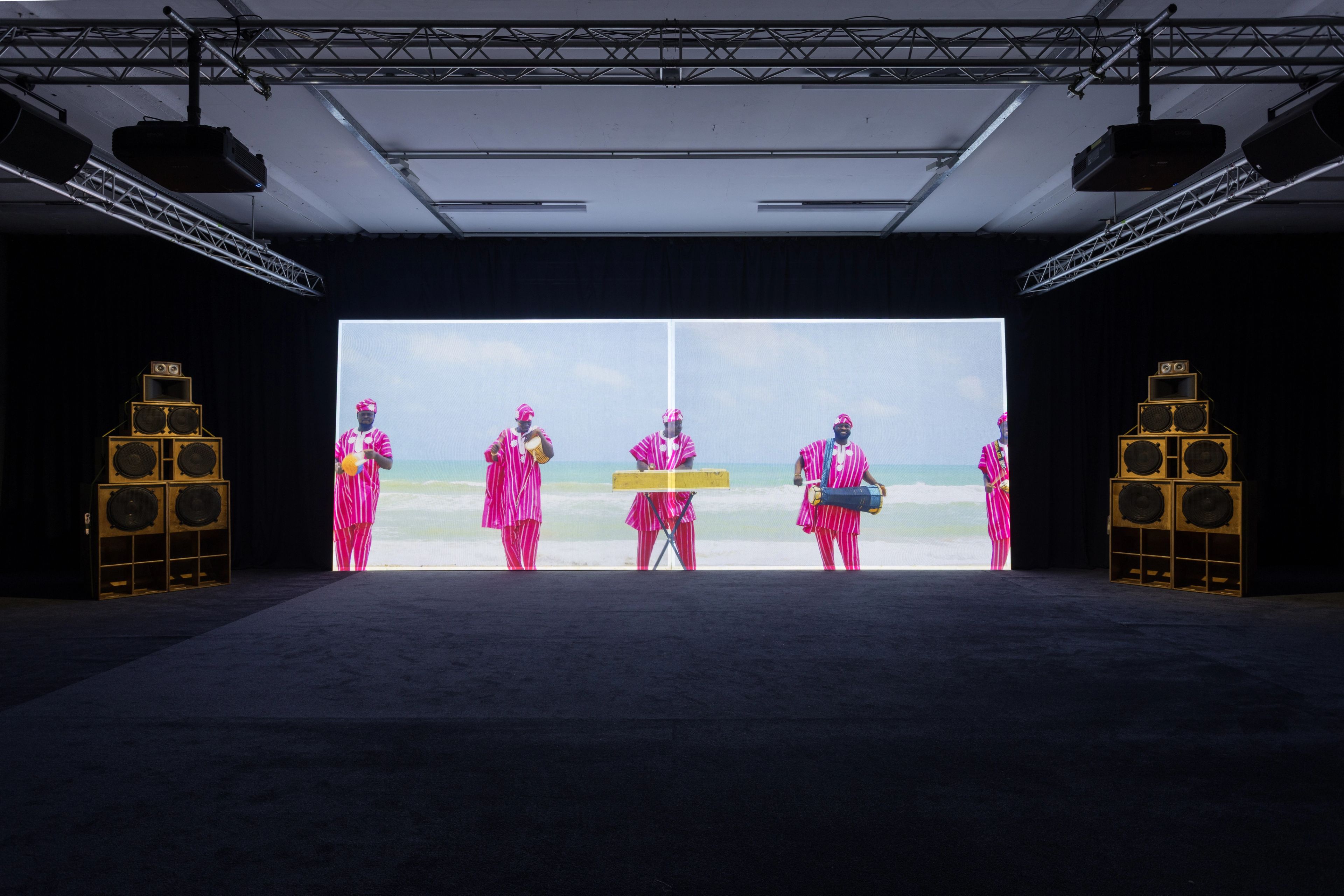A dark exhibition room with a large video screen showing people in pink on a beach, flanked by two tall stacks of wooden speakers.