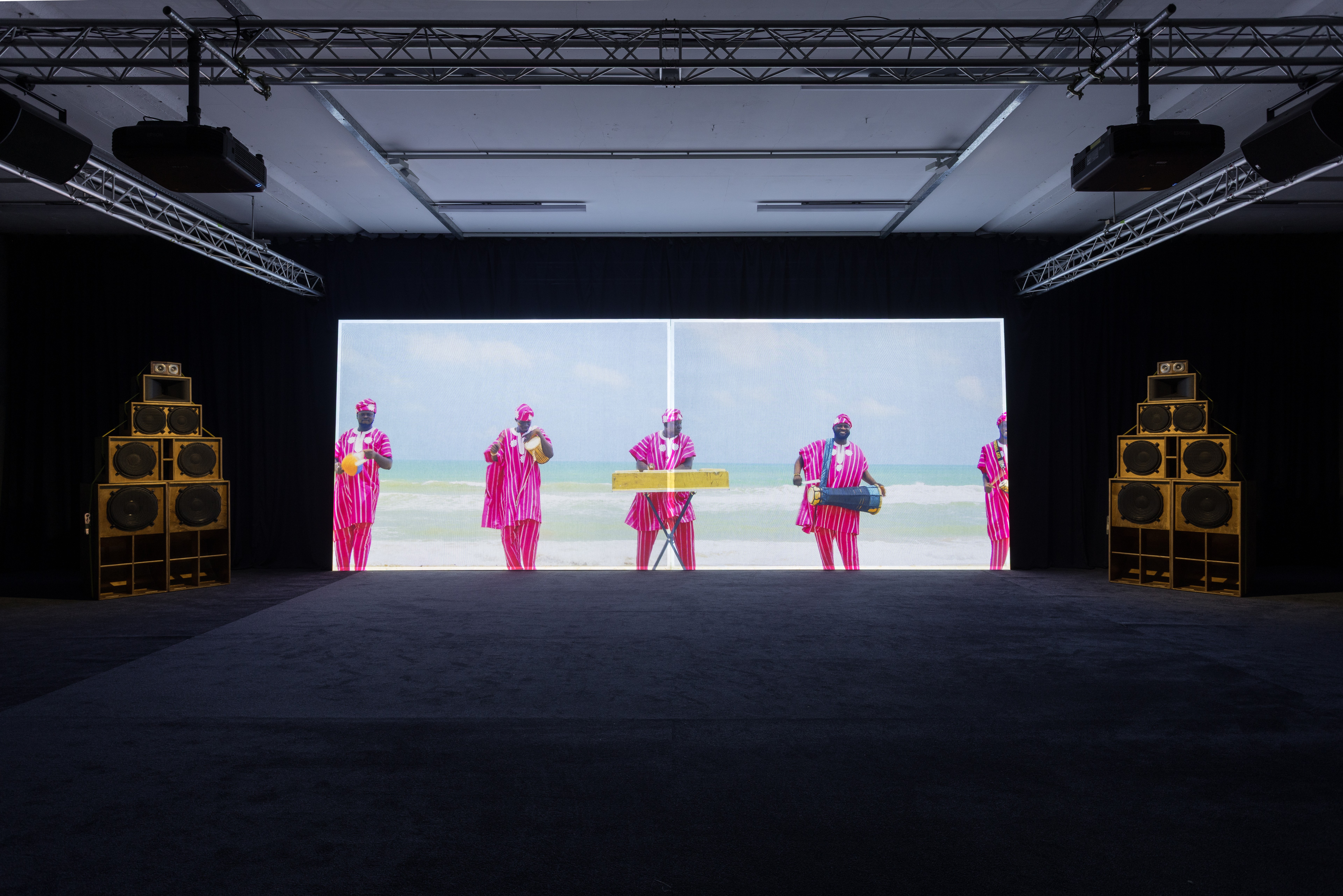 A dark exhibition room with a large video screen showing people in pink on a beach, flanked by two tall stacks of wooden speakers.