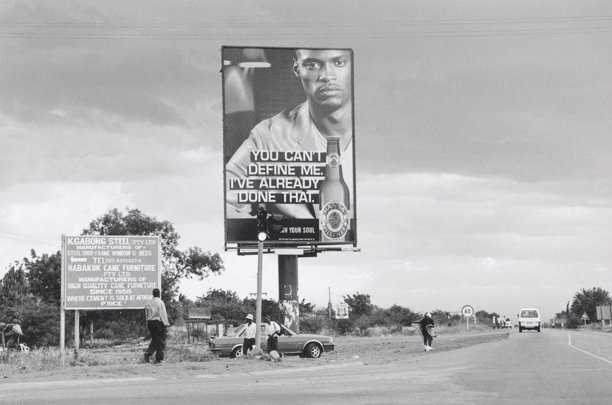 Black and white roadside scene with a large billboard featuring a man and beer bottle with the text "YOU CAN'T DEFINE ME. I'VE ALREADY DONE THAT.", a smaller sign, people, and a road.