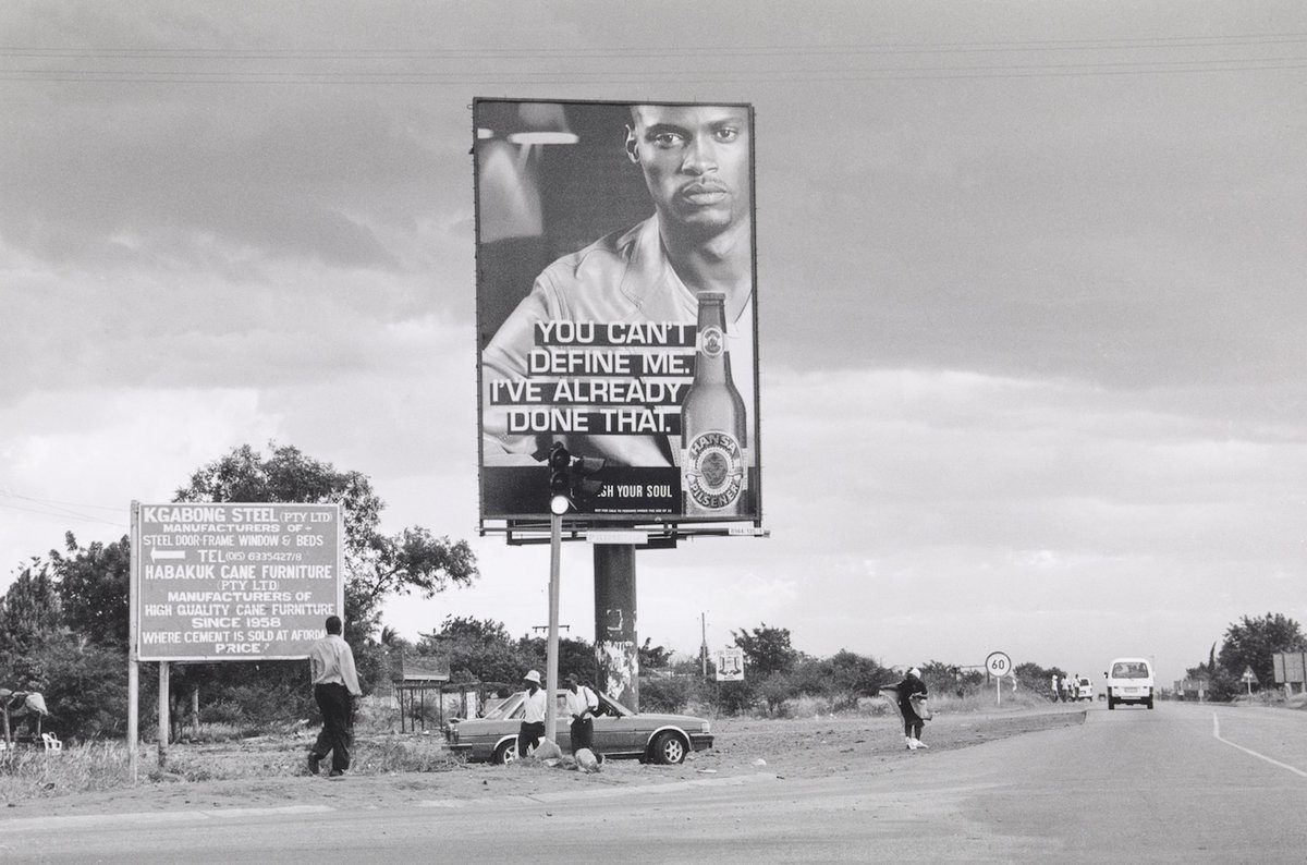 Black and white roadside scene with a large billboard featuring a man and beer bottle with the text "YOU CAN'T DEFINE ME. I'VE ALREADY DONE THAT.", a smaller sign, people, and a road.