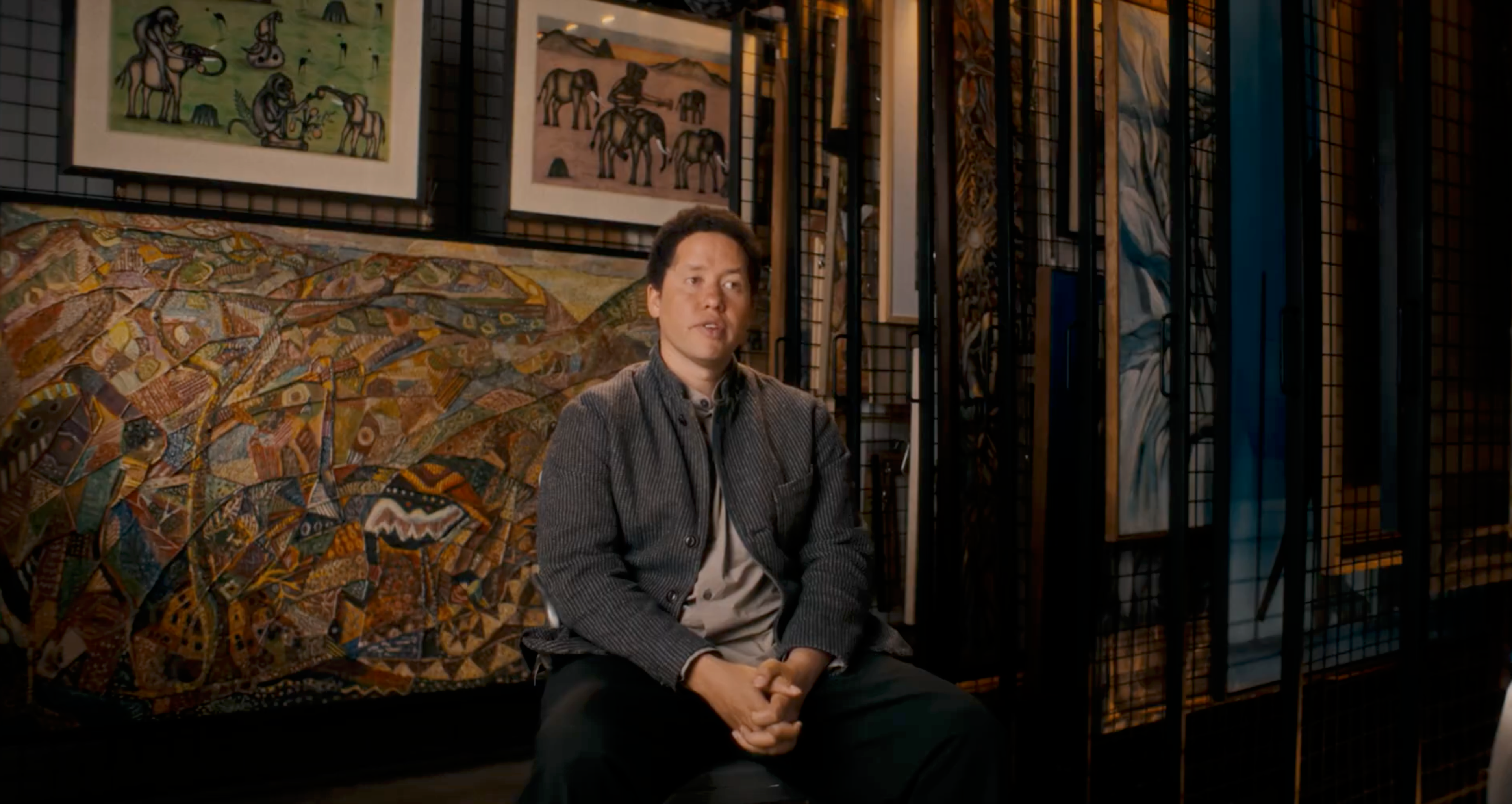 A man sits in an art gallery, a large colorful abstract painting behind him and two smaller elephant paintings above.