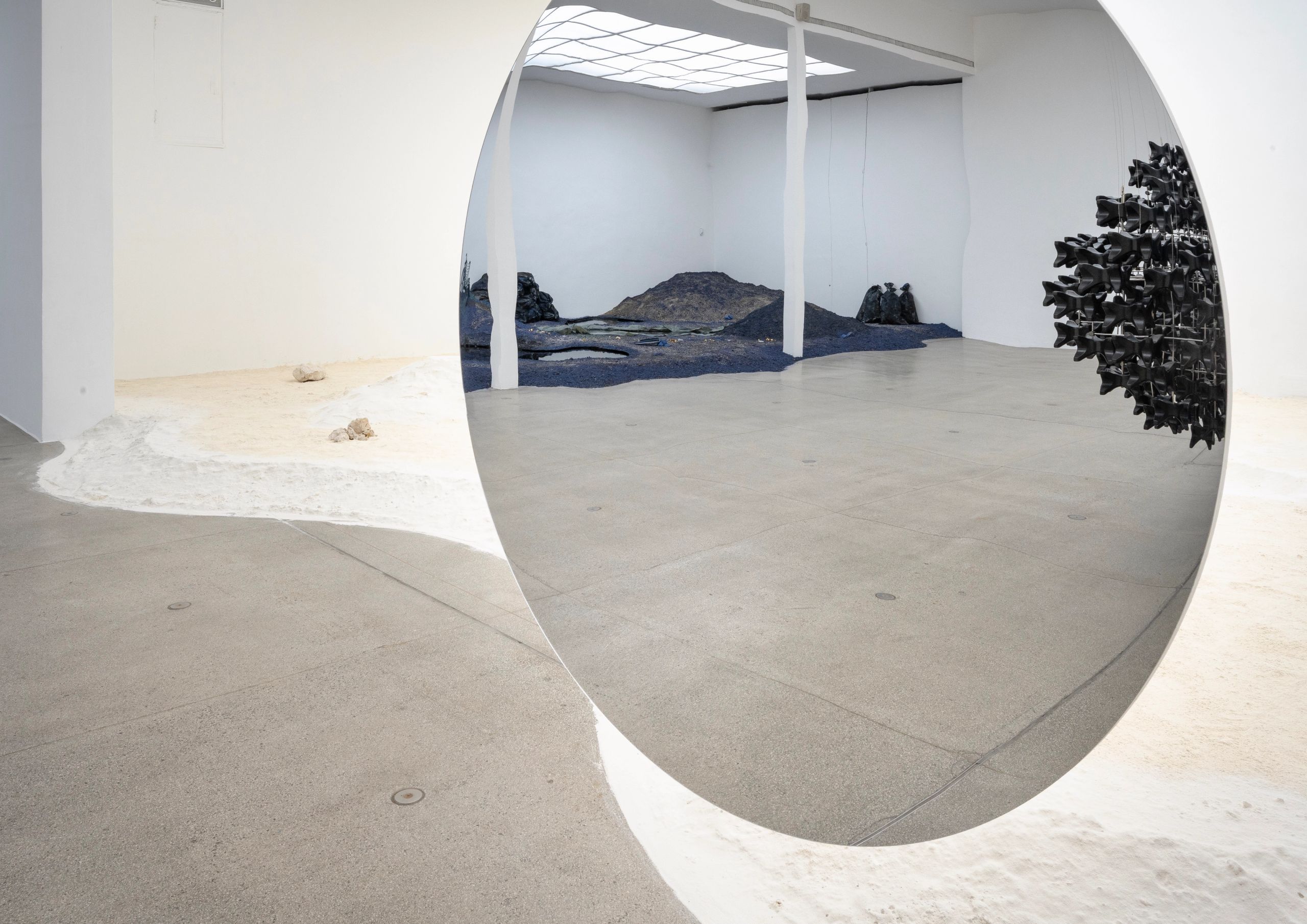 A large circular mirror reflects a dark, sculptural landscape within an art gallery, while the foreground shows a white wavy material and rocks on the floor.