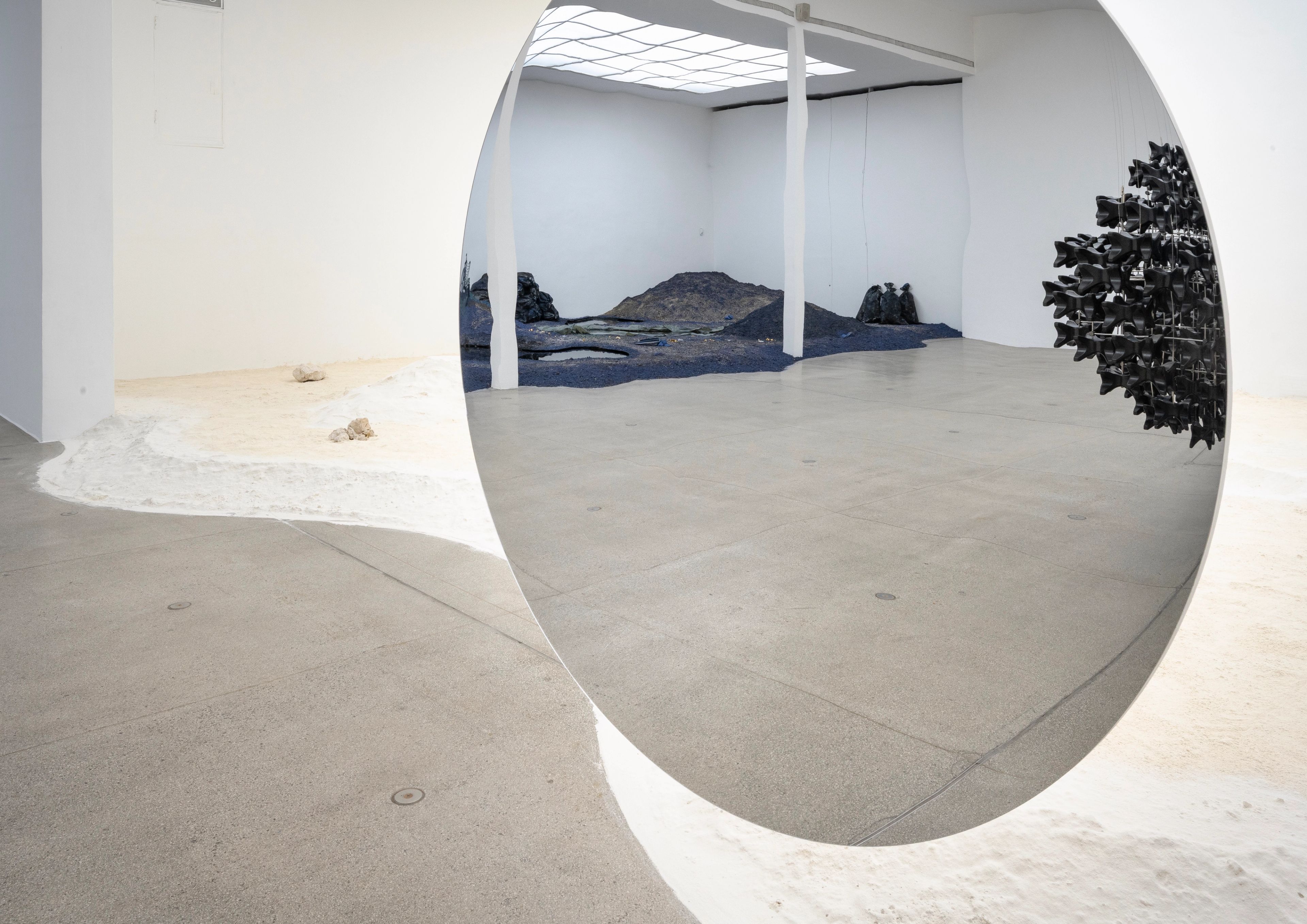 A large circular mirror reflects a dark, sculptural landscape within an art gallery, while the foreground shows a white wavy material and rocks on the floor.