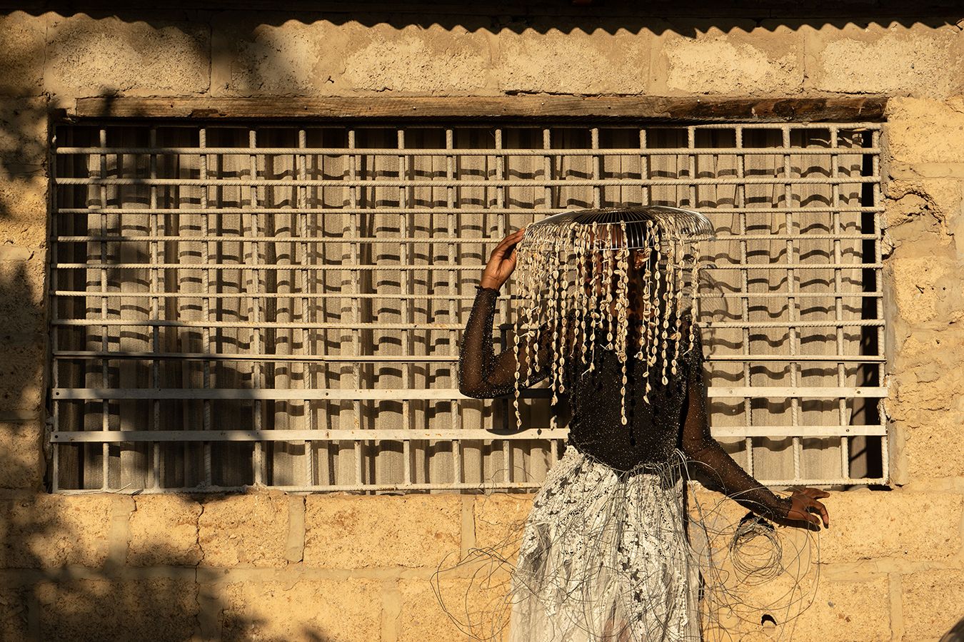 A figure in a black top, white textured skirt, and an elaborate beaded headpiece obscuring their face stands before a barred window and a stone wall.