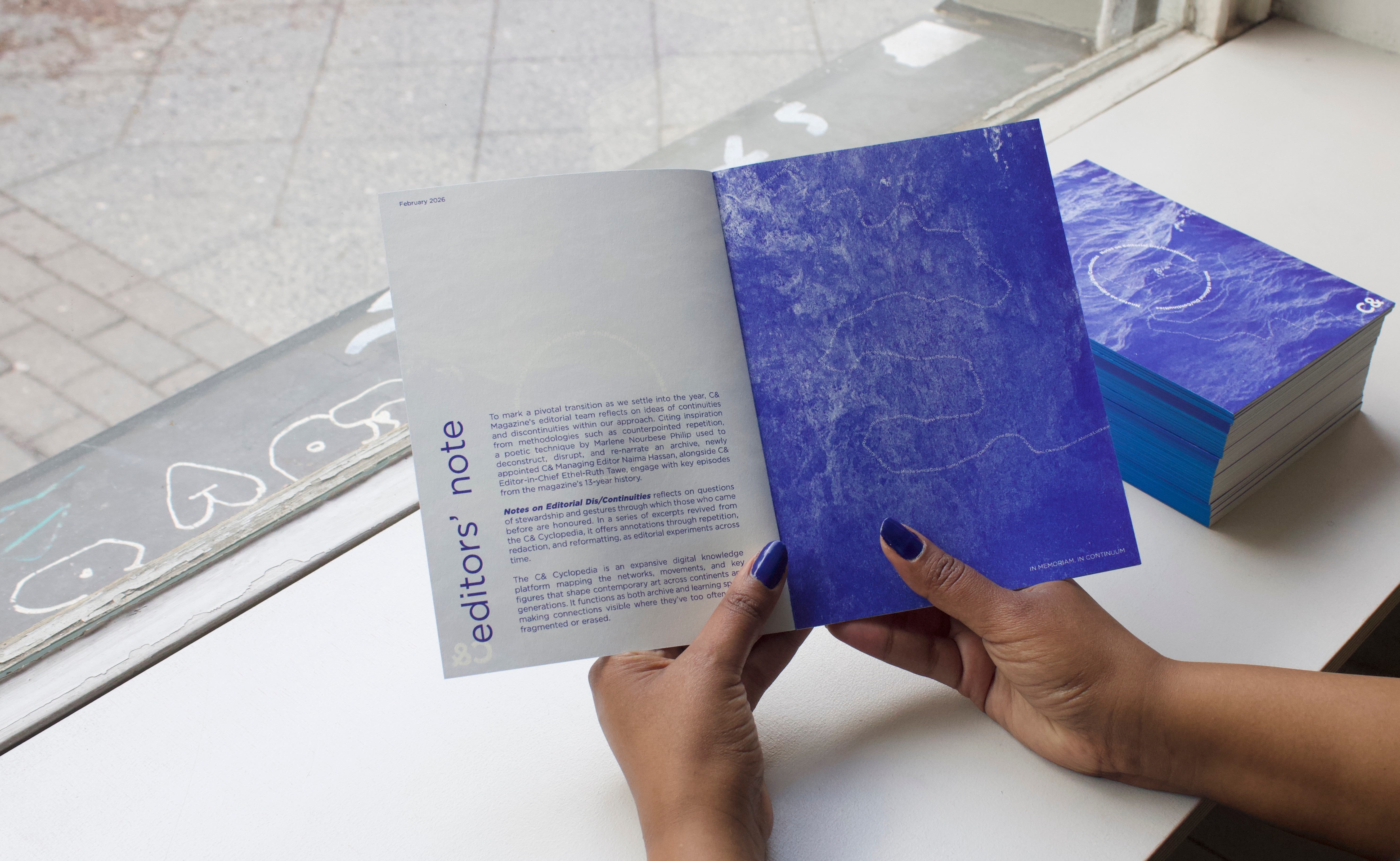 Hands hold an open publication with an "editors' note" page and a blue page featuring a faint abstract design, with a stack of similar booklets nearby.