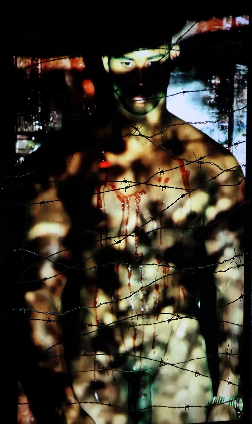 A dark, intense image of a shirtless man with blood dripping down his chest, partially obscured by strands of barbed wire.