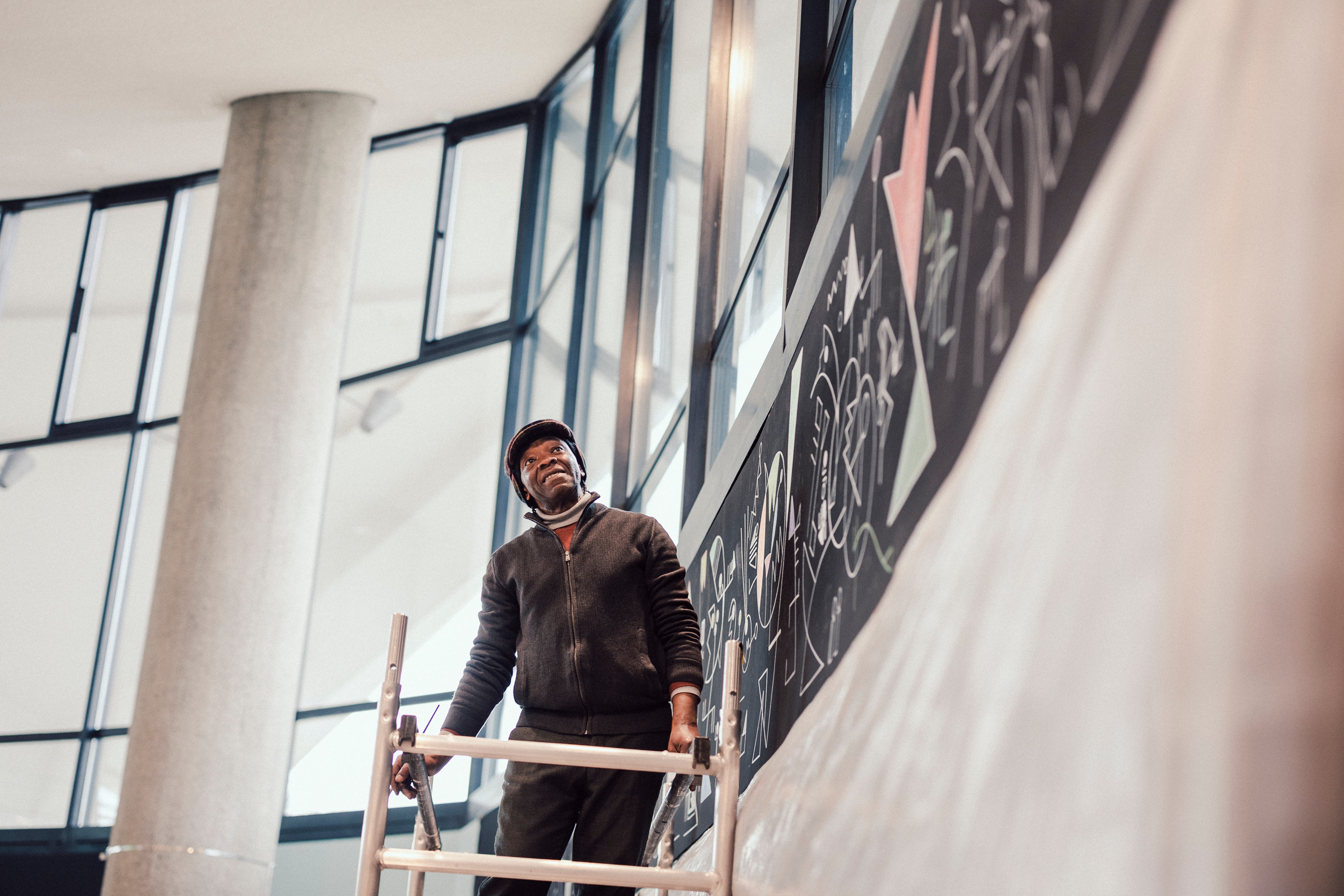 Smiling Black man on a ladder looking up at a mural with chalk drawings.