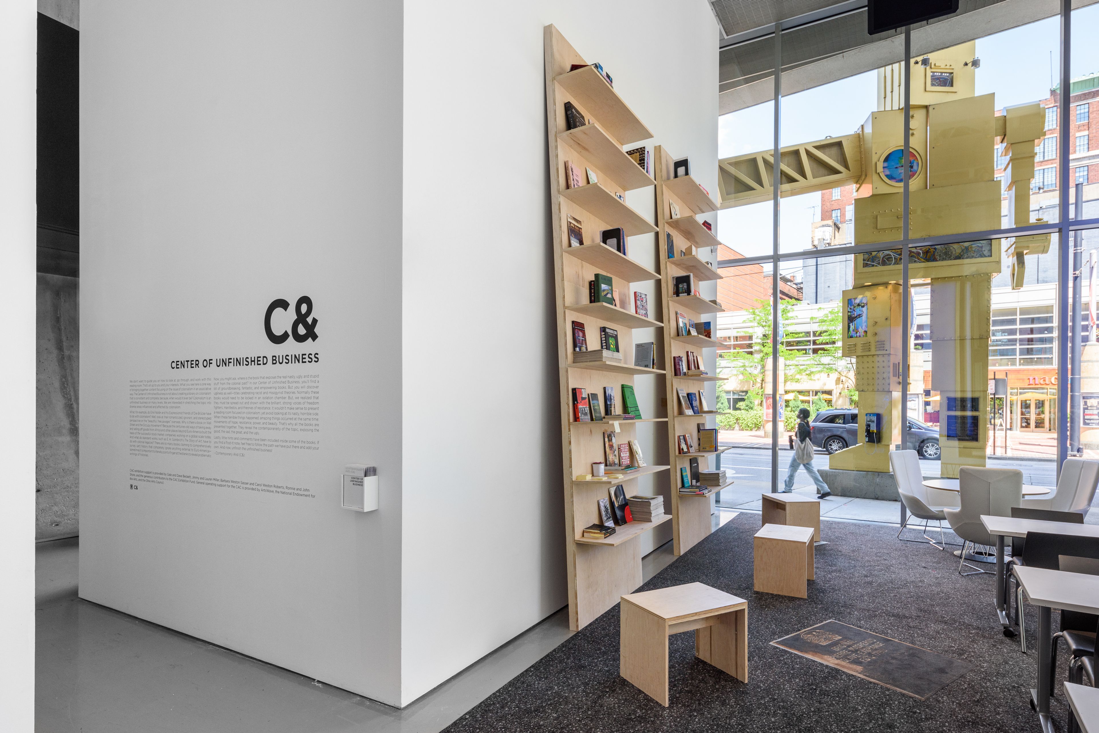 A modern interior features a white wall with "Center of Unfinished Business" text, a tall wooden bookshelf, minimalist seating, and a large window revealing a city street with a prominent yellow robot sculpture.