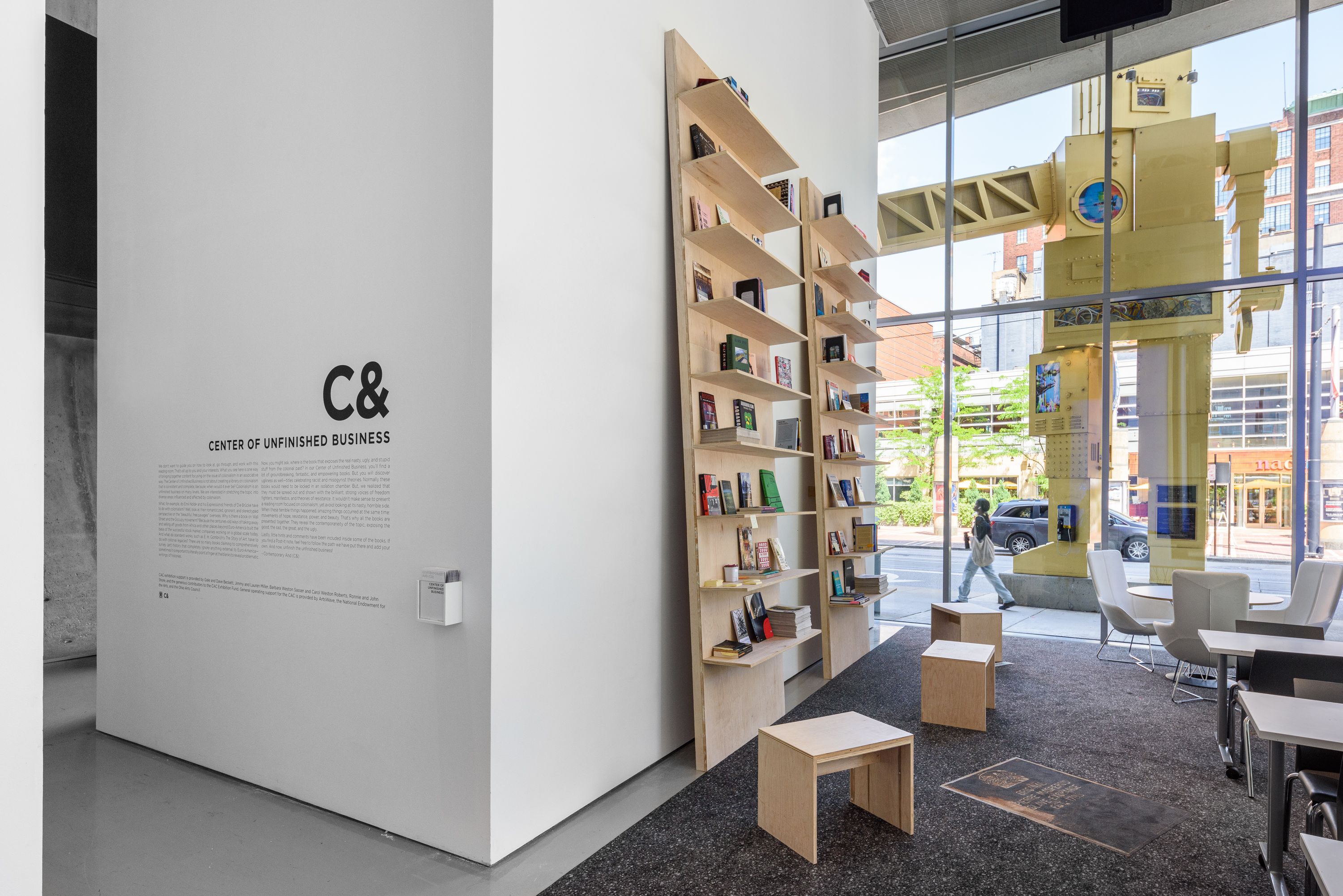 A modern interior features a white wall with "Center of Unfinished Business" text, a tall wooden bookshelf, minimalist seating, and a large window revealing a city street with a prominent yellow robot sculpture.
