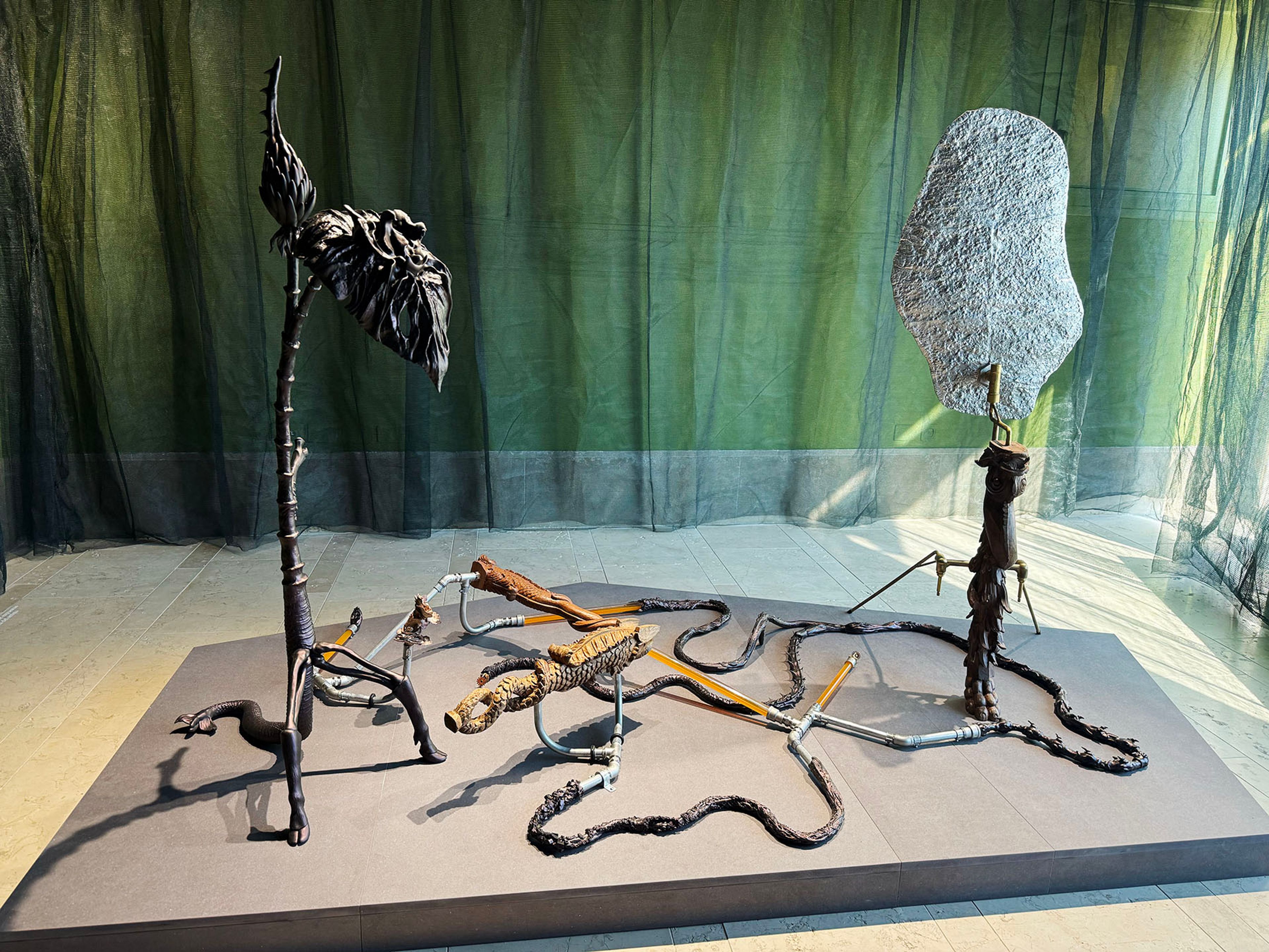 An art installation featuring two dark, organic, vertical sculptures, one with a textured gray oval top and the other with a dark leaf-like form, interconnected by winding pipes and textured elements on a grey platform.