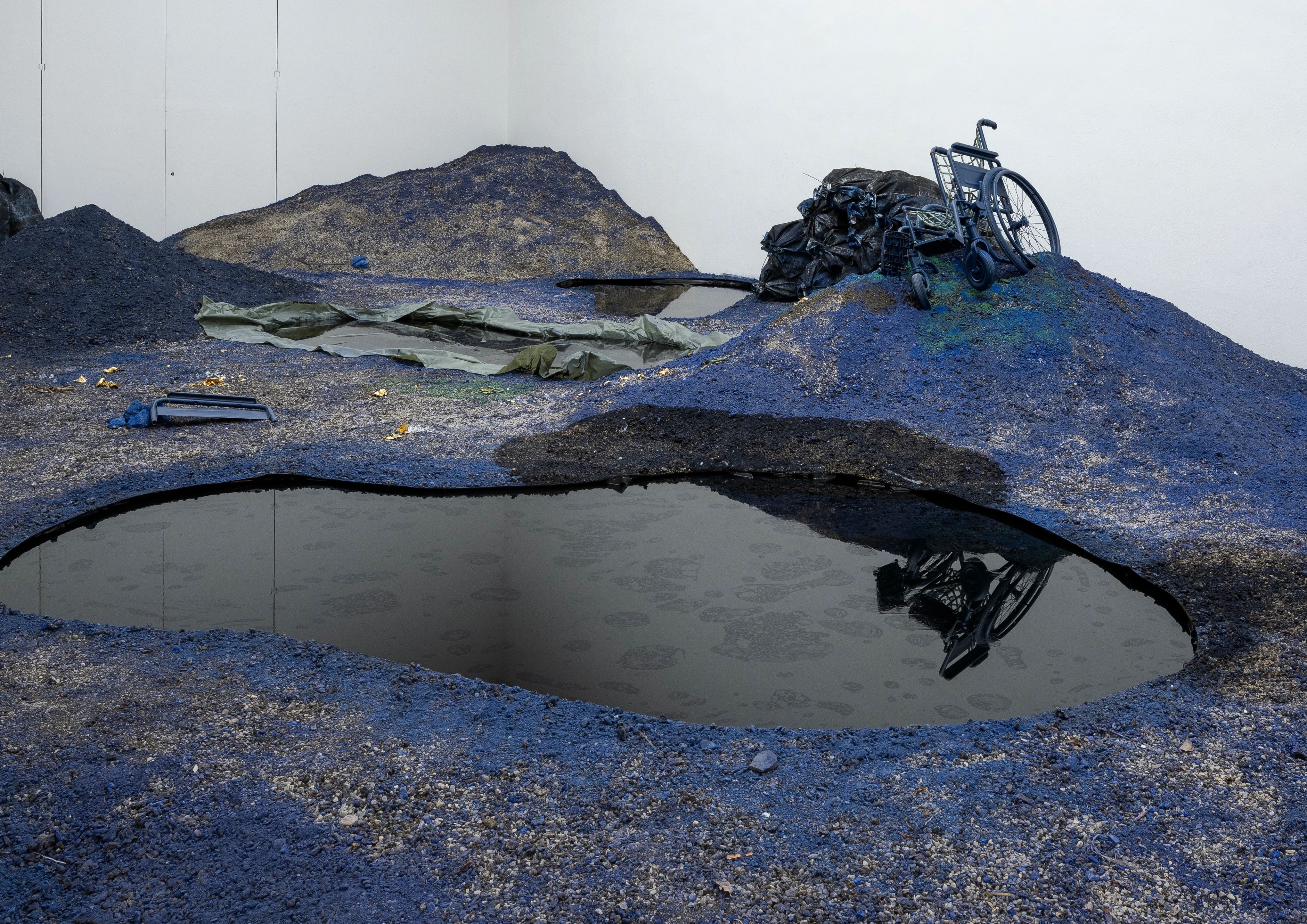 Art installation with blue and black mounds, dark pools, and a crumpled blue wheelchair reflected in the water.