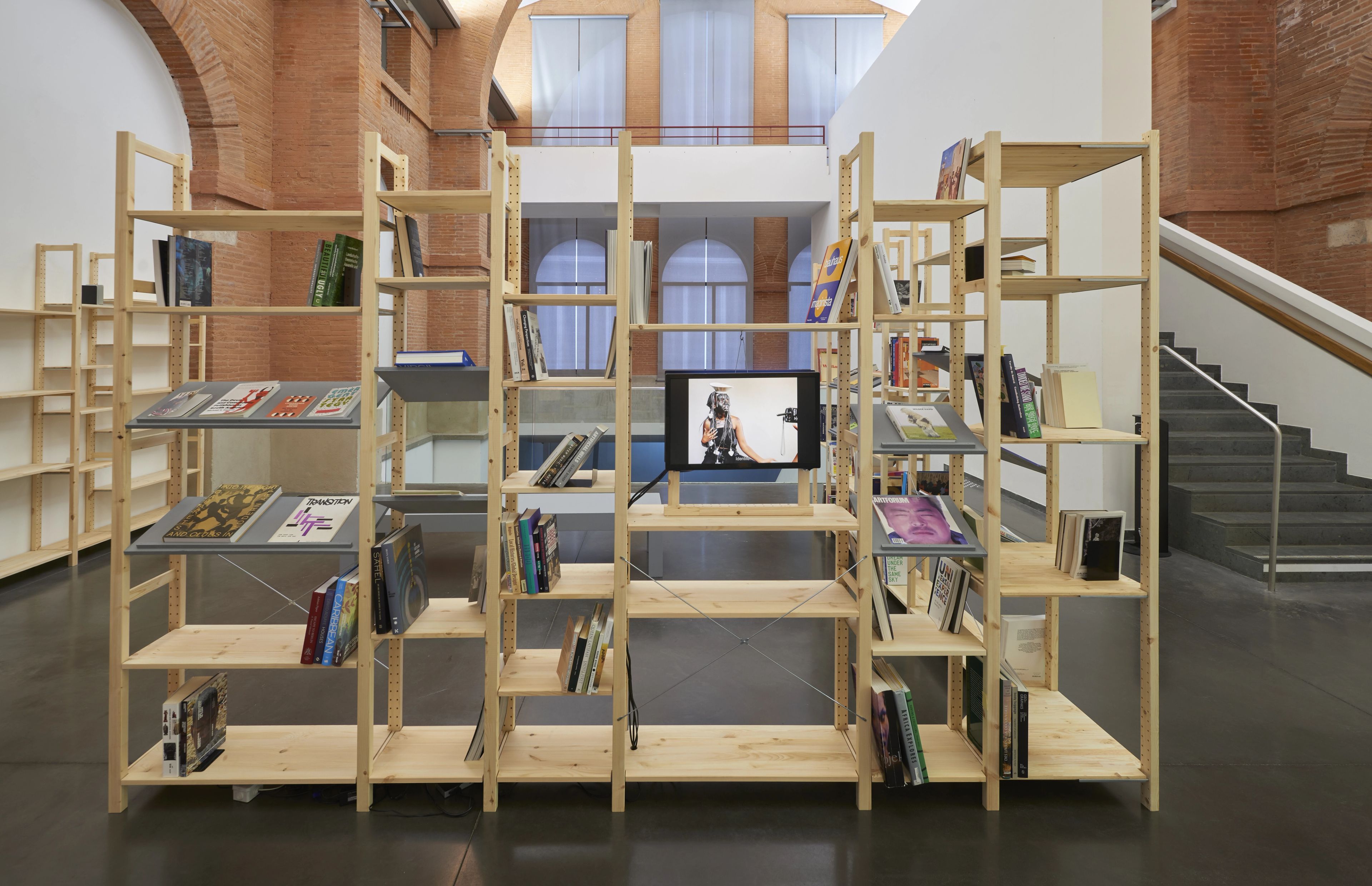 A light wood modular shelving system displays books, magazines, and a monitor playing a video of a person, set in a spacious building with brick walls and a staircase.