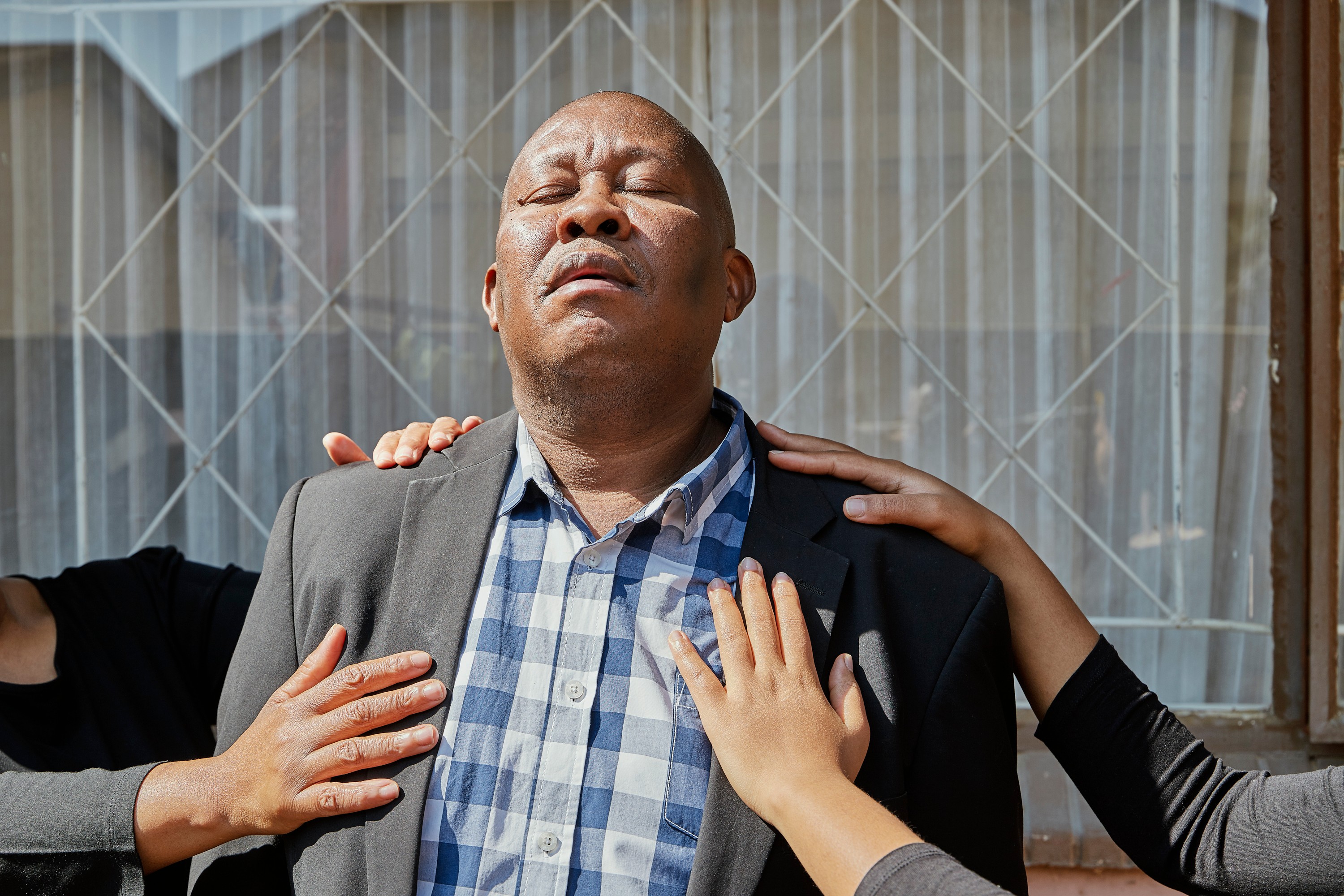 A man with closed eyes and head tilted up, with multiple hands resting on his chest and shoulders.