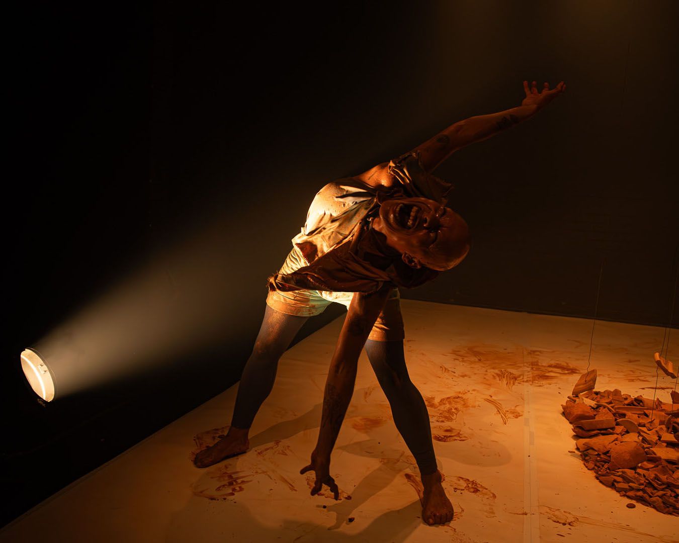 A performer in a spotlight, leaning back intensely with mouth open, on a red-stained stage next to a pile of broken clay.