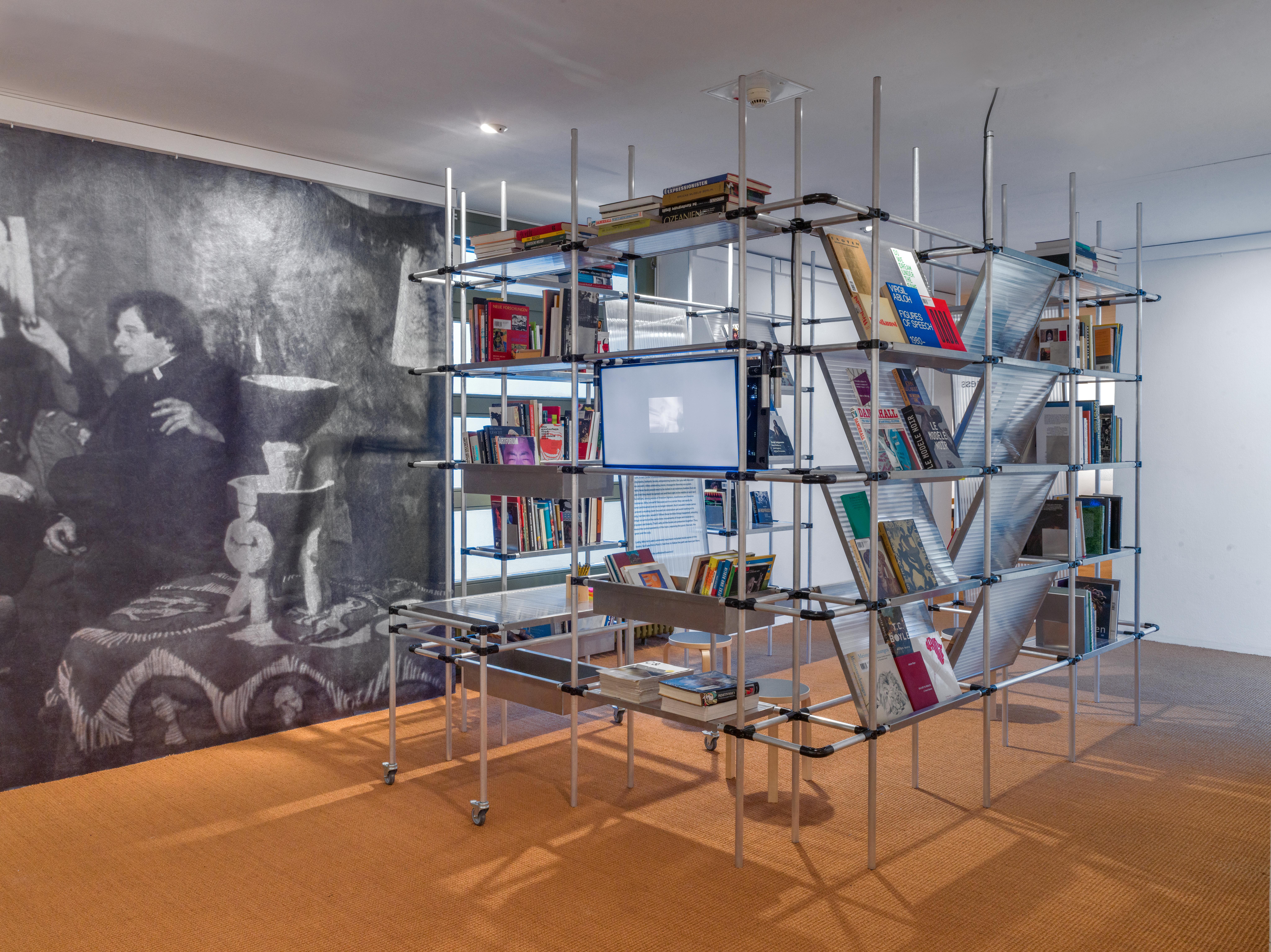 A modern, multi-level metal pipe bookshelf filled with books and a monitor, stands in front of a large black-and-white mural of people.