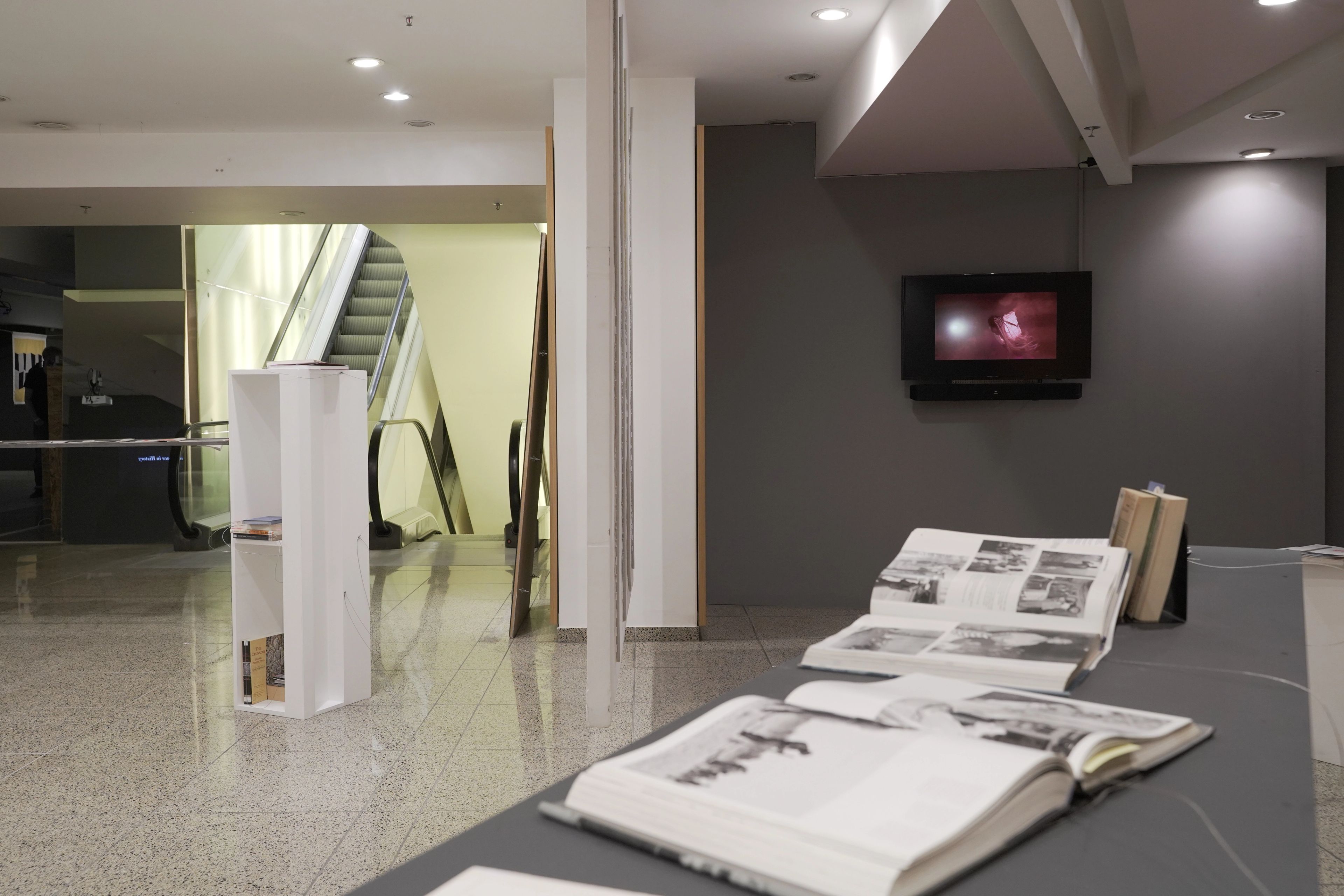 Open books with photos on a display table in a modern interior with a TV screen on a grey wall and escalators in the background.