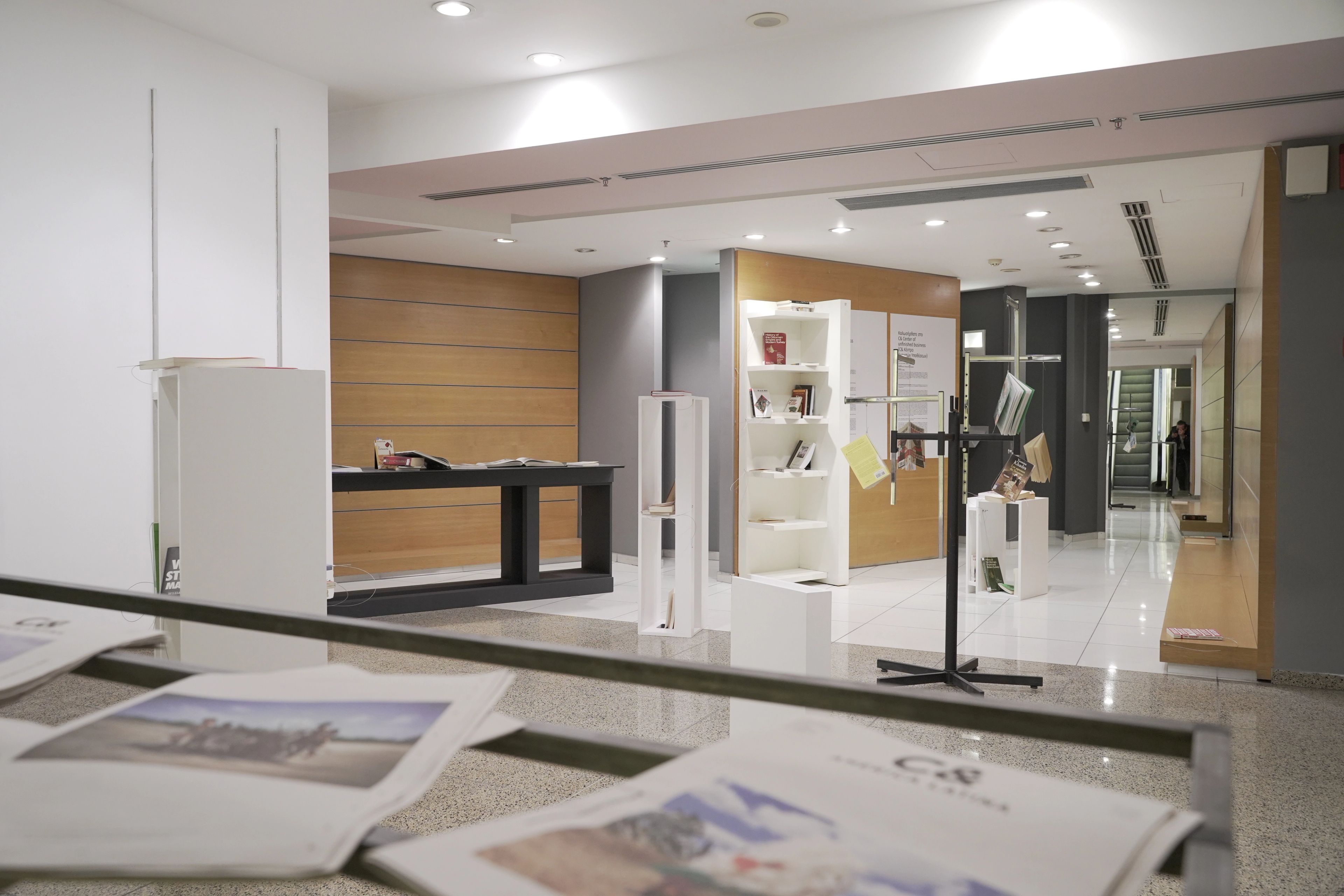 A bright, modern gallery or lobby space with white display shelves, tables, and magazine racks. Magazines are visible in the blurry foreground.