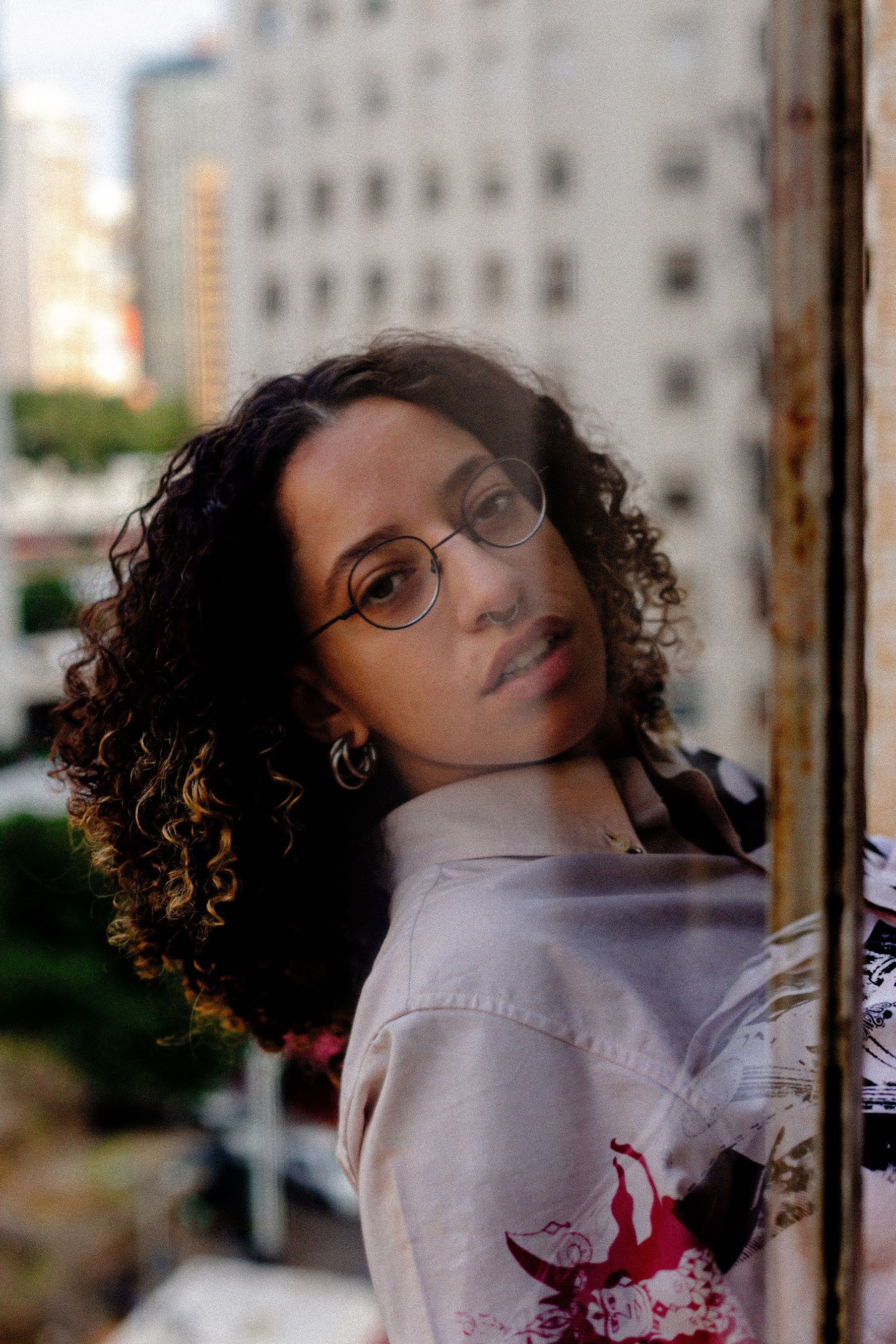 A young woman with curly hair, round glasses, and a nose ring looks directly at the camera, her face partly shadowed, against a blurred city background.