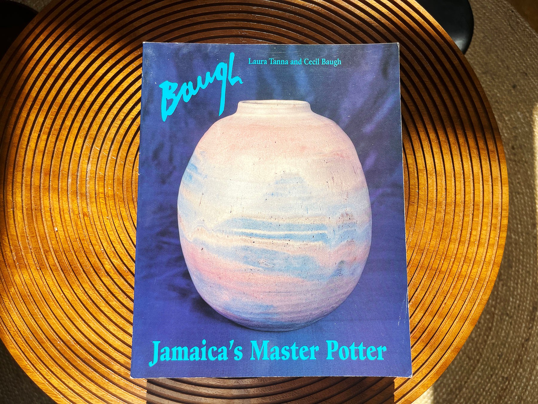 Book "Baugh: Jamaica's Master Potter" with a pink and blue vase on the cover, resting on a wooden table.