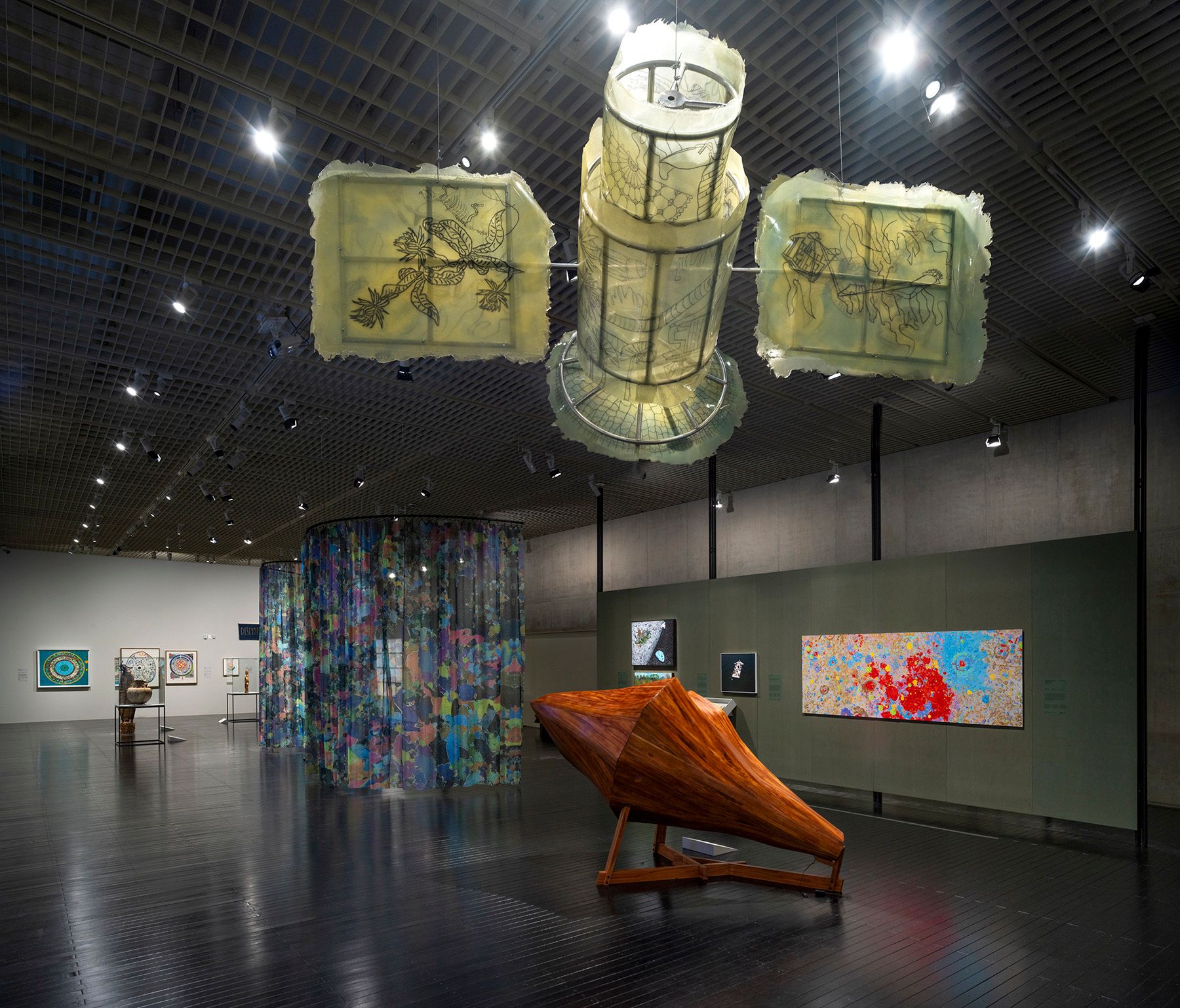 A modern art gallery showcasing various artworks, including large translucent ceiling sculptures, a wooden boat-shaped sculpture, and colorful abstract pieces on walls and a cylindrical curtain.
