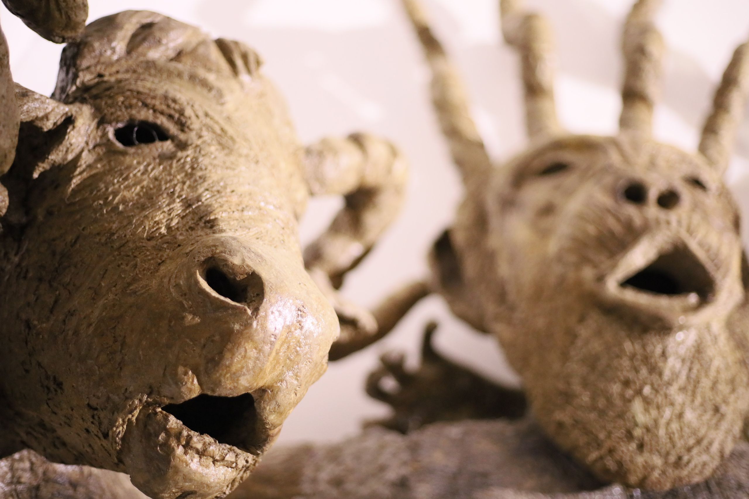 Two textured tan ceramic sculptures of open-mouthed creature heads; one ram-like in focus, one blurry with multiple protrusions.