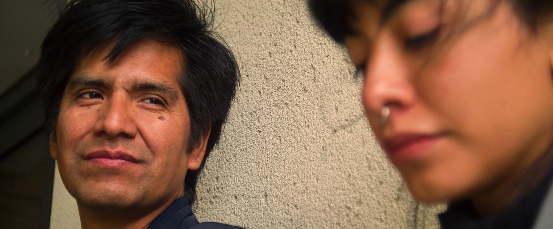 Close-up of a smiling man with dark hair, with a blurry person wearing a nose ring partially visible on the right.
