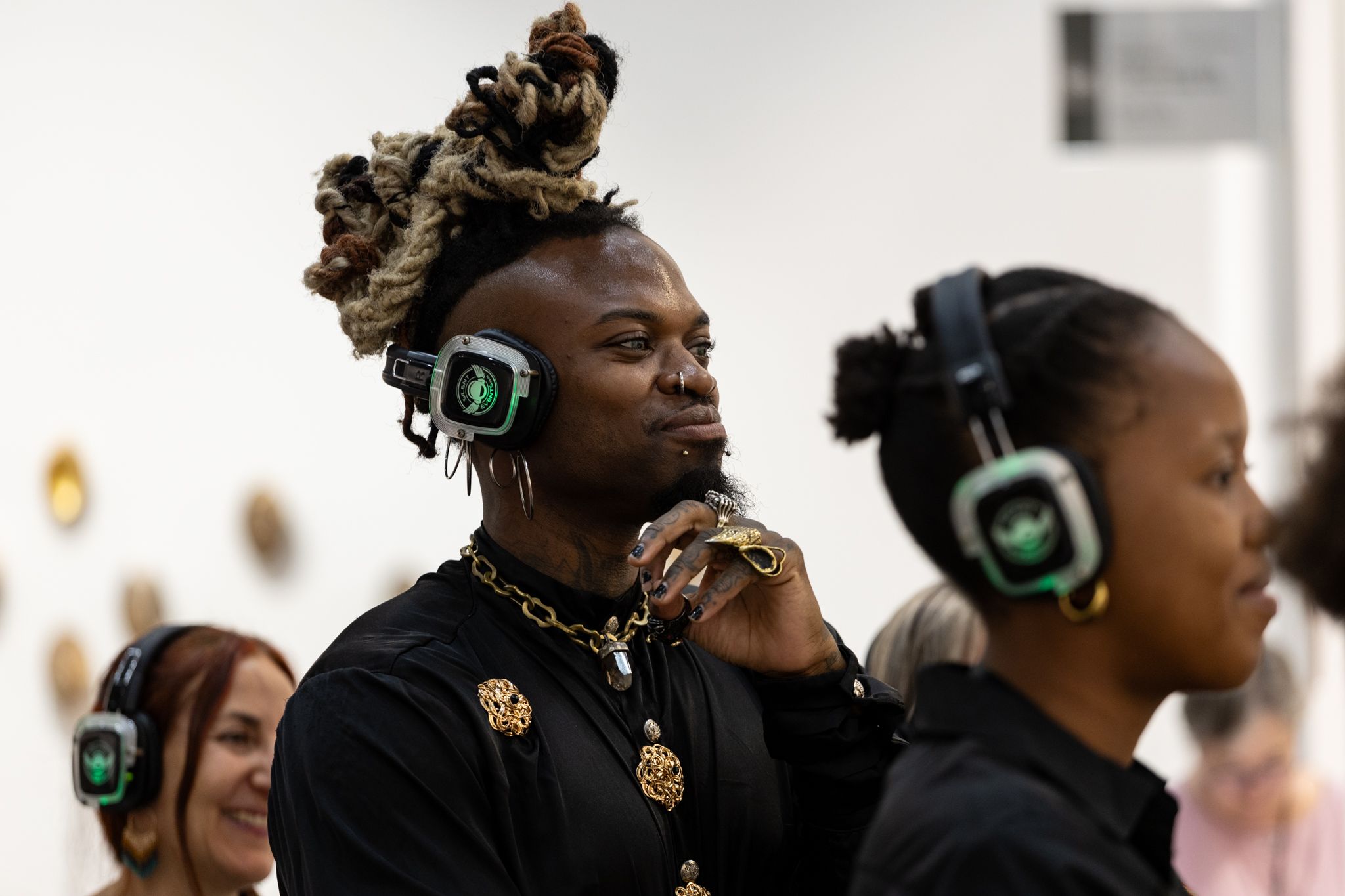 A Black person with dreadlocks, facial piercings, and intricate jewelry smiles while wearing headphones, with other people also wearing headphones nearby.