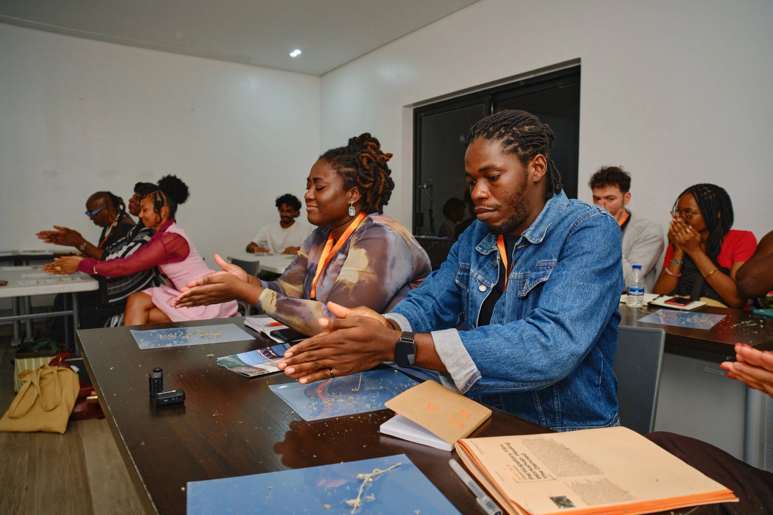 A group of people sit at tables in a classroom, many with their hands open and palms facing up.