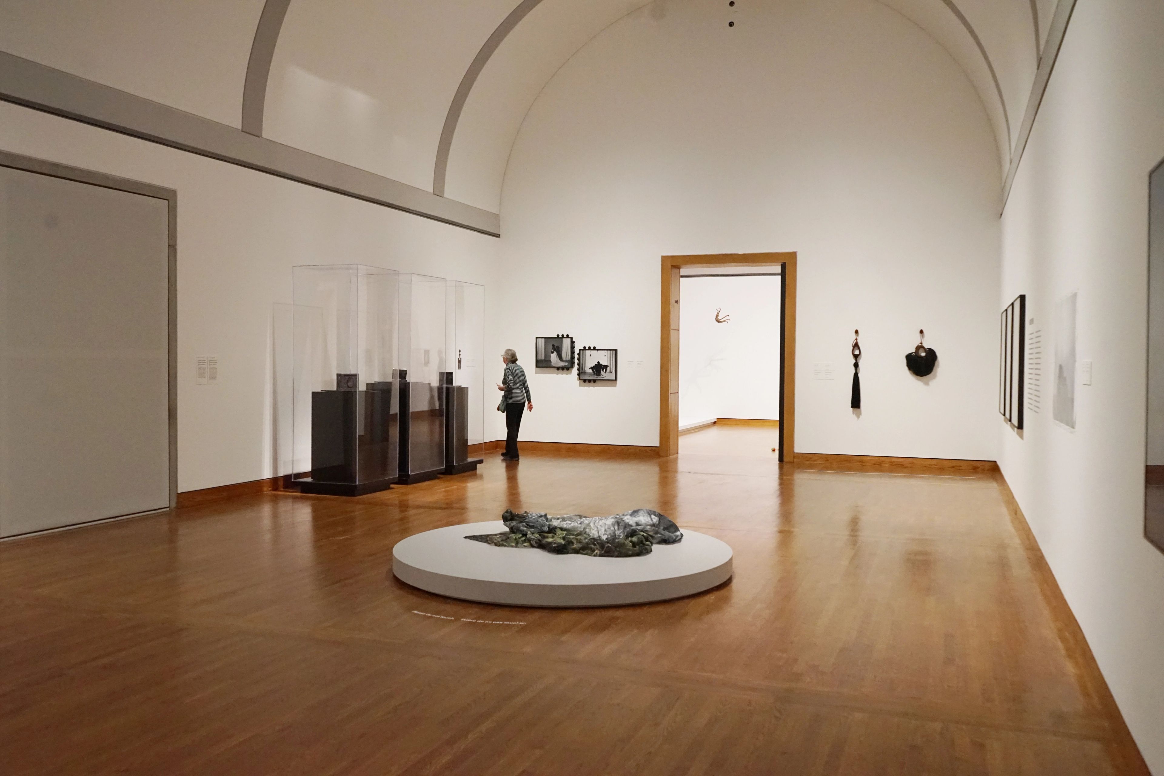 A well-lit museum gallery with white walls, wooden floor, and a vaulted ceiling, featuring a sculpture on a central platform, framed art, and a person viewing displays.