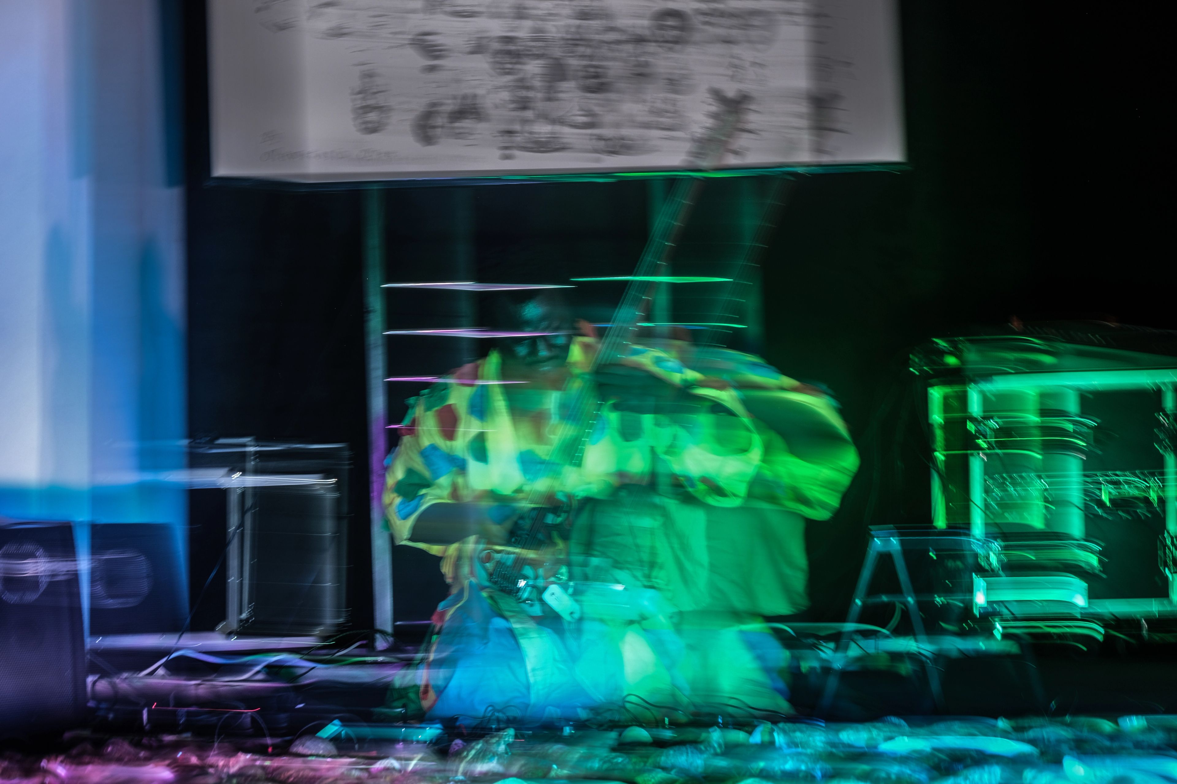 Motion-blurred figure in a colorful costume on a stage with vibrant green and blue lighting.