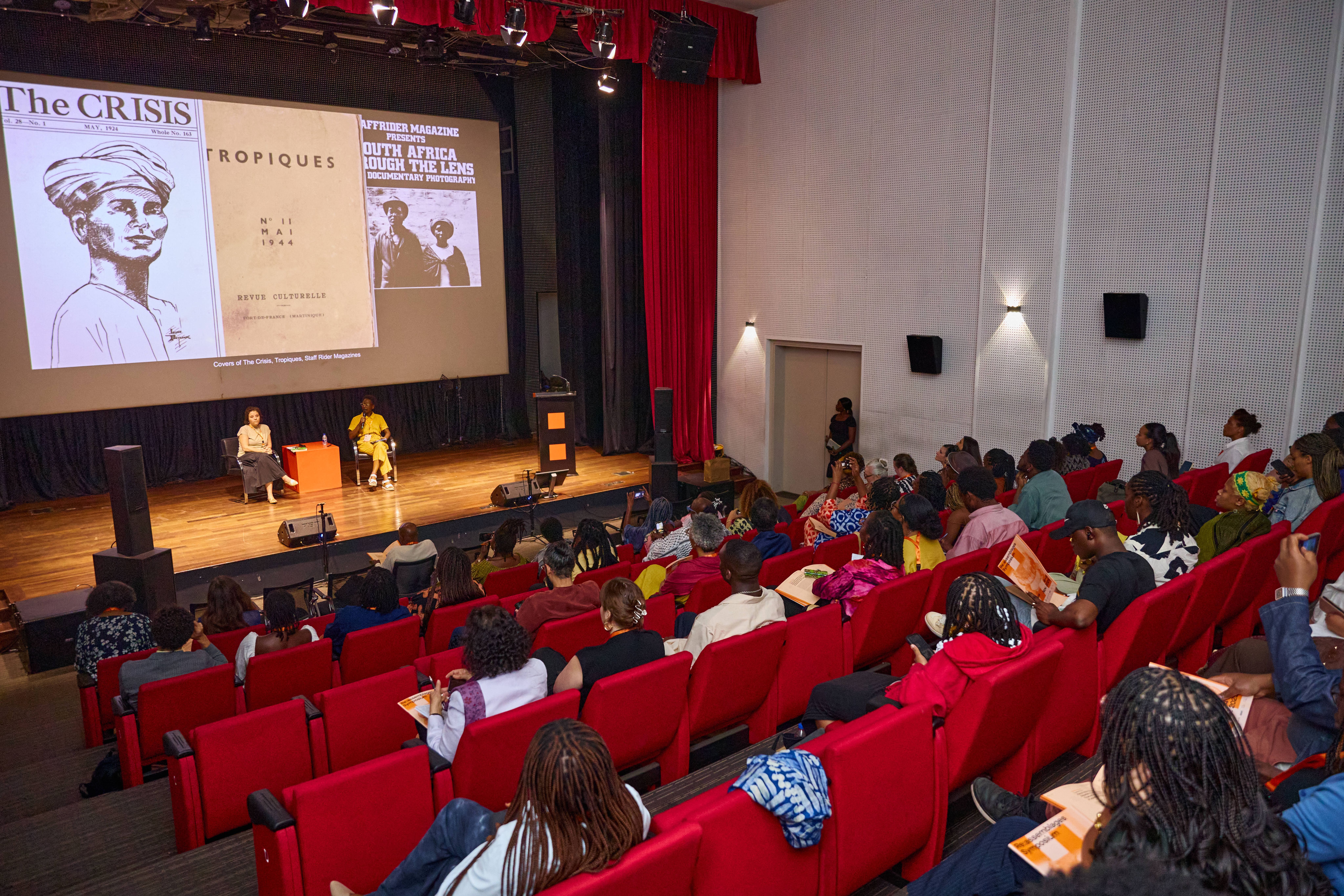 Audience in an auditorium watching two speakers on a stage with historical magazine covers projected on a large screen.