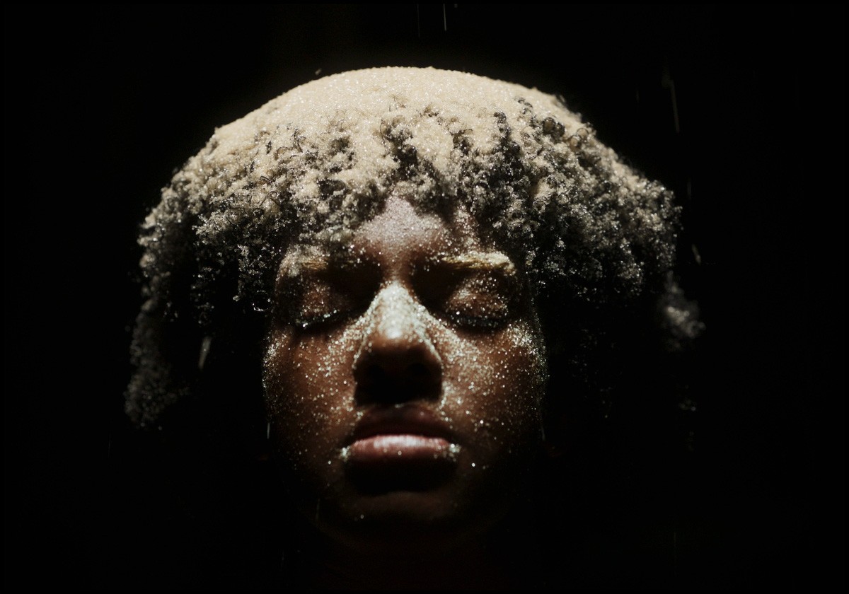 A person's face and hair are covered in white sugar-like granules, with closed eyes and dripping liquid.